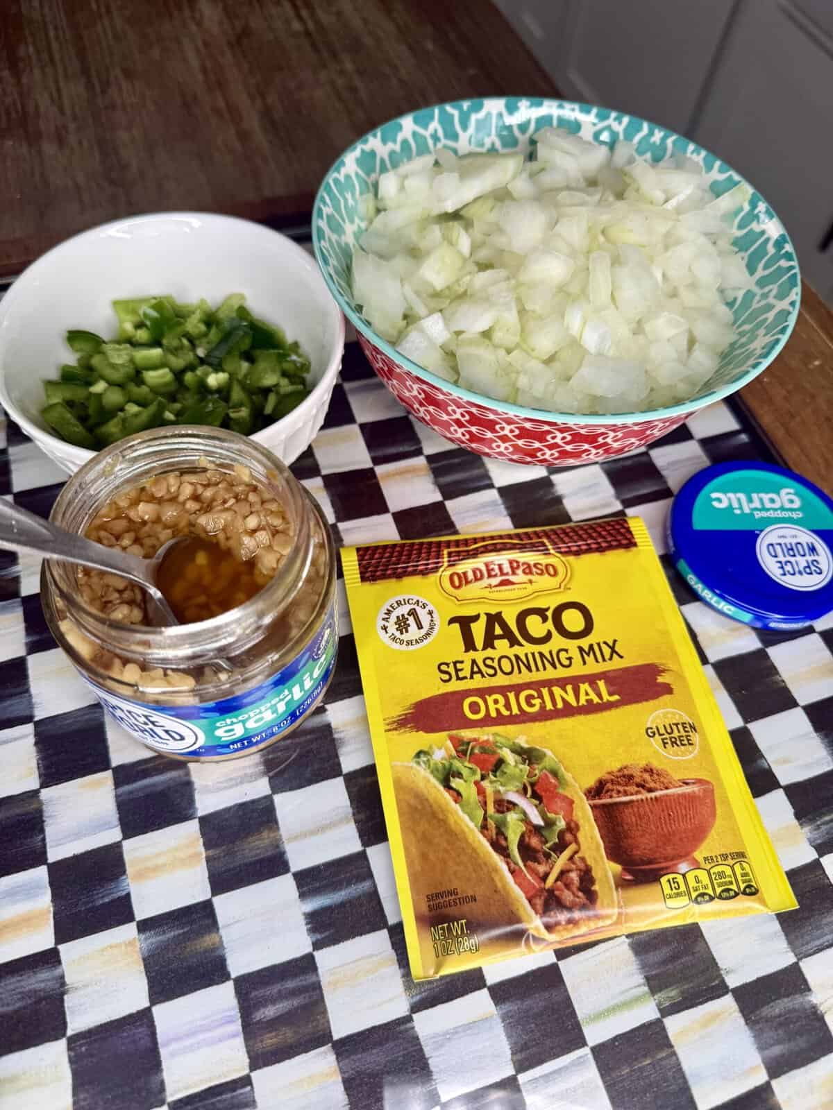 Chopped onions, chopped green peppers, jar of minced garlic, taco seasoning mix, and a blue garlic lid are arranged on a checkered surface next to a bowl and kitchen counter.