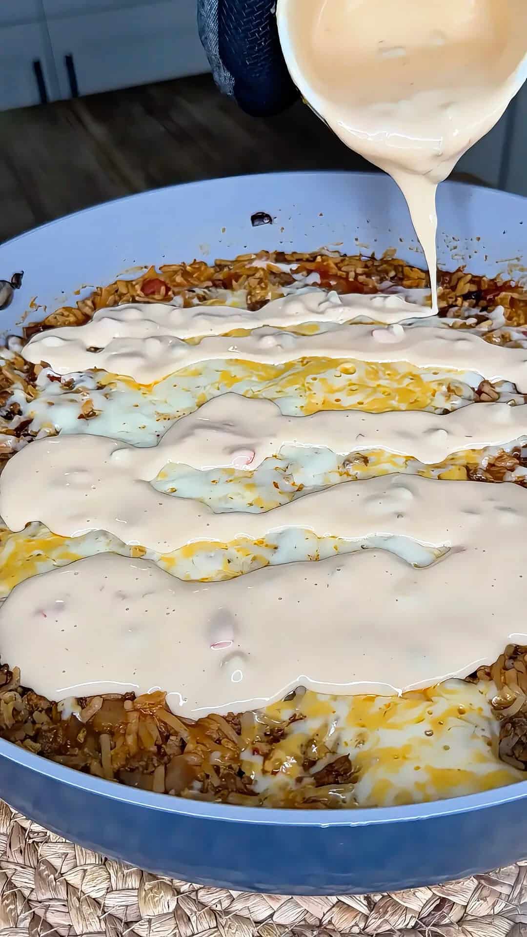A creamy sauce is being poured over a skillet filled with rice, ground meat, cheese, and vegetables. The mixture is partially melted and still cooking in the pan.