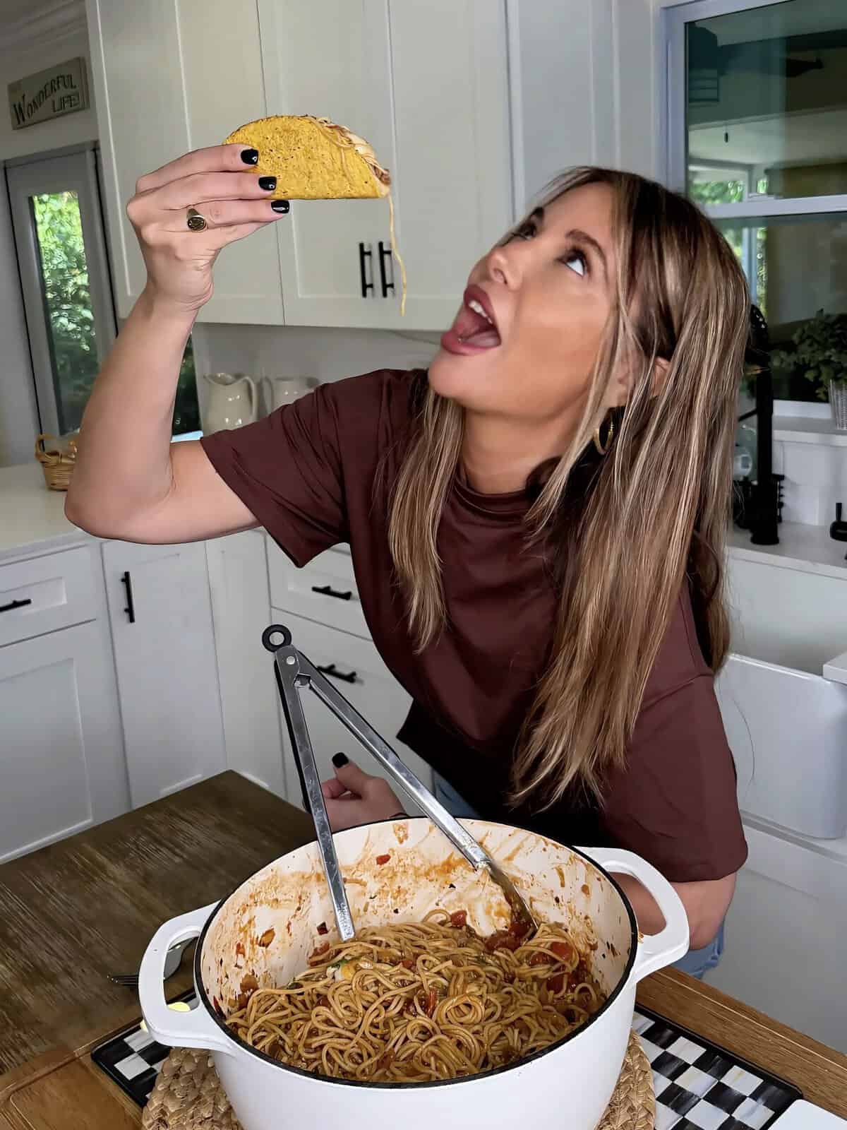 A woman in a kitchen holds up a taco filled with spaghetti, looking up with her mouth open to take a bite. A pot of spaghetti is on the counter in front of her.