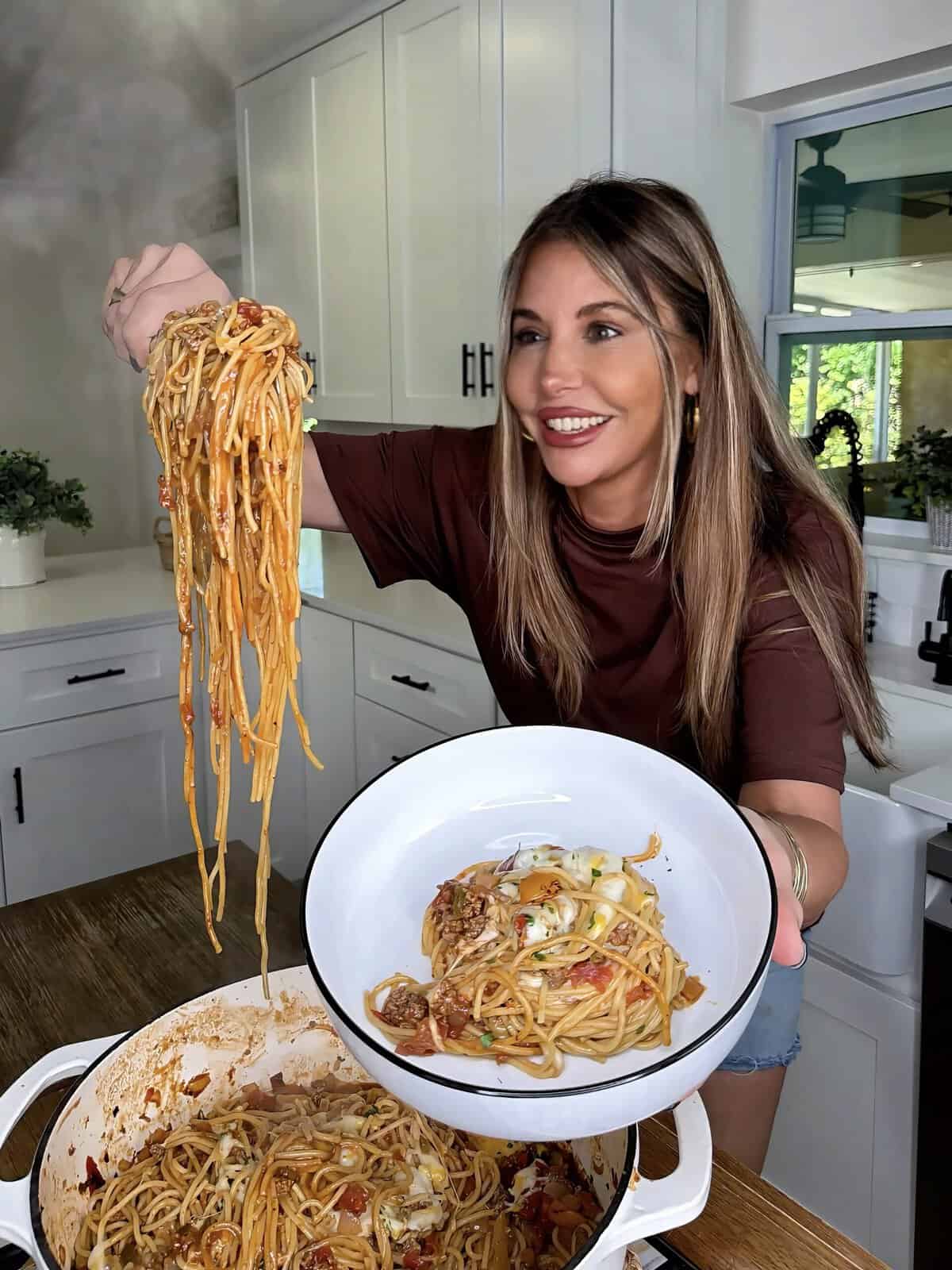 A smiling woman in a kitchen serves a large portion of spaghetti with sauce from a pot into a white bowl, holding both spaghetti and bowl toward the camera.