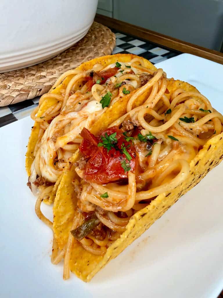 Two hard taco shells filled with spaghetti, tomato sauce, ground meat, melted cheese, and garnished with chopped herbs, placed on a white plate.