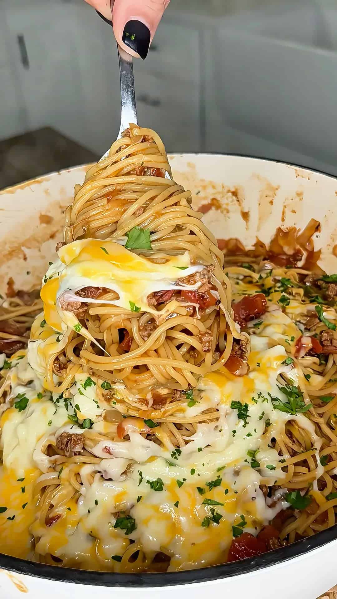 A fork lifts a serving of cheesy spaghetti with meat sauce from a pot. Melted cheese and herbs are sprinkled on top, and the dish looks hot and freshly made.