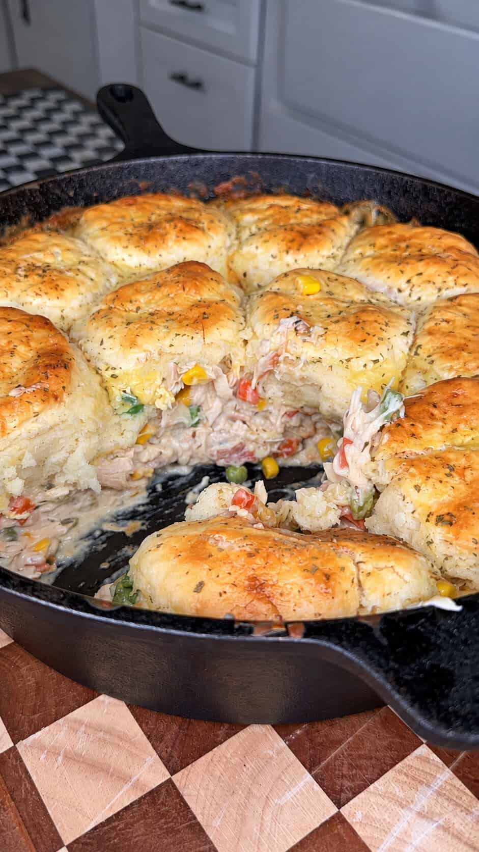 A cast-iron skillet filled with golden, flaky biscuits baked on top of a creamy chicken and vegetable mixture, with a portion removed to reveal the filling.