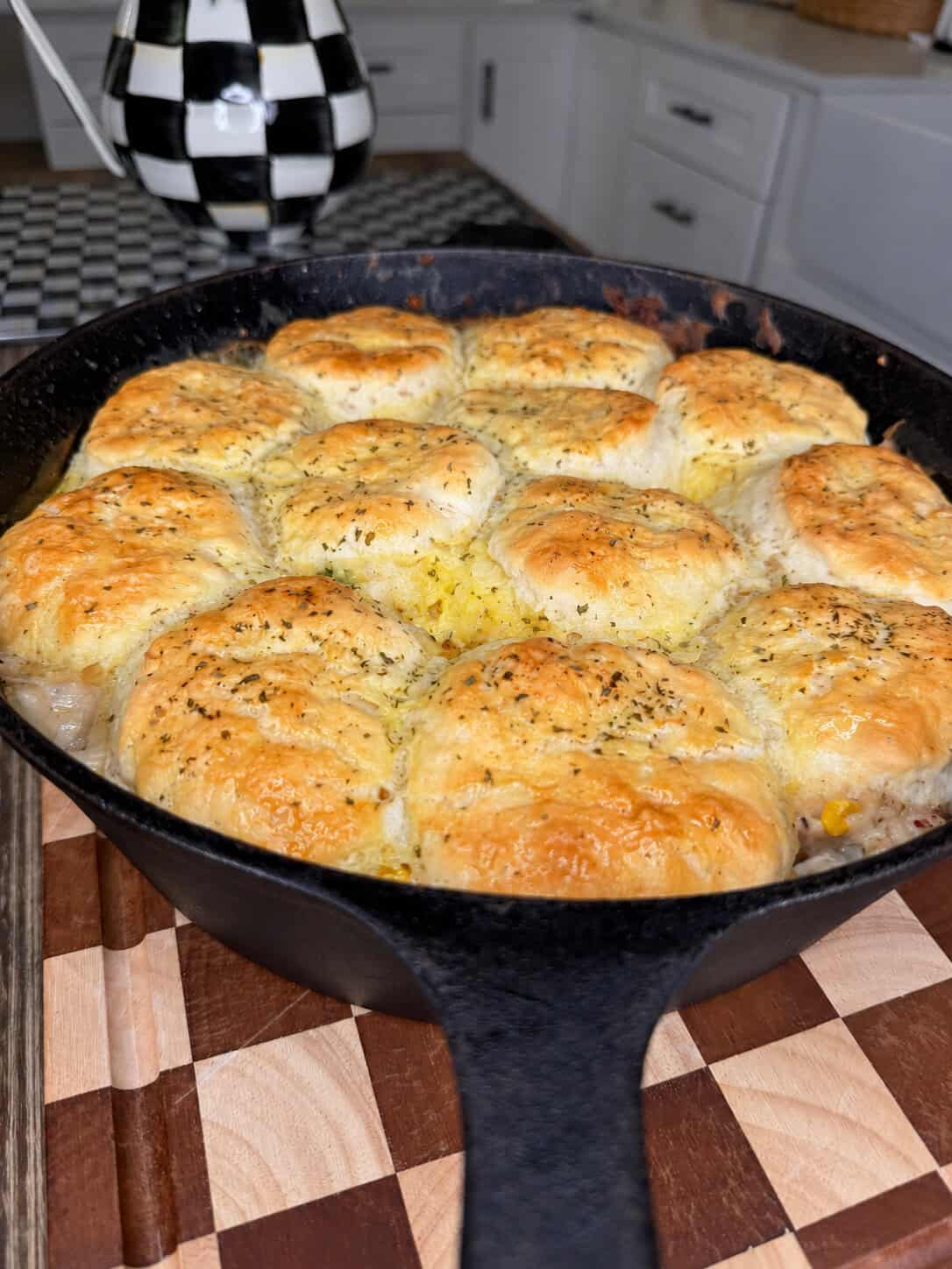 A cast iron skillet filled with golden, herb-topped biscuits baked on top of a savory casserole, resting on a wooden cutting board in a kitchen setting.