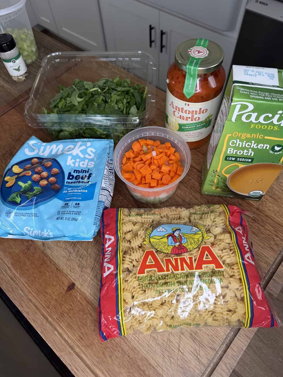 Ingredients on a wooden counter: frozen mini beef meatballs, chopped carrots, spinach, jar of pasta sauce, chicken broth carton, and a bag of rotini pasta.
