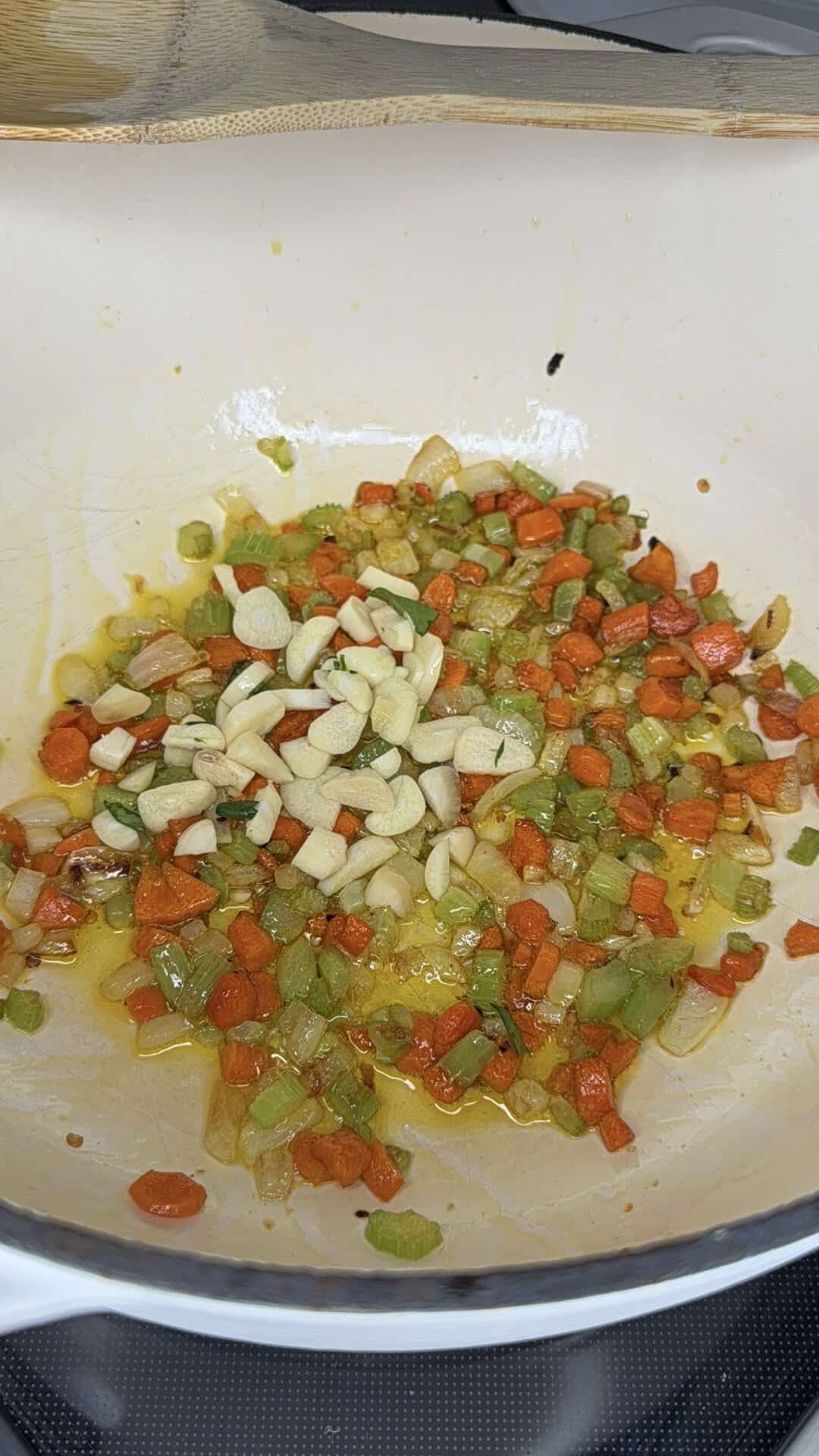 Chopped carrots, celery, onions, and sliced garlic are sautéing in oil in a white pot on a stovetop.