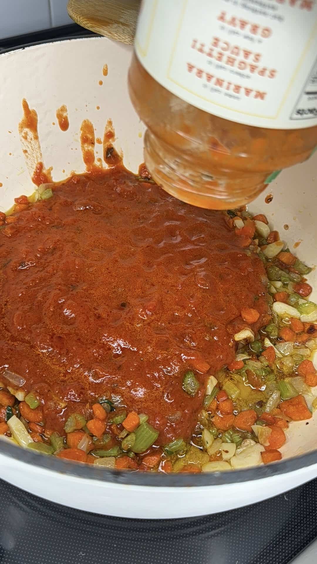 A jar of tomato sauce is being poured over chopped carrots, celery, and onions in a white pot on a stovetop.