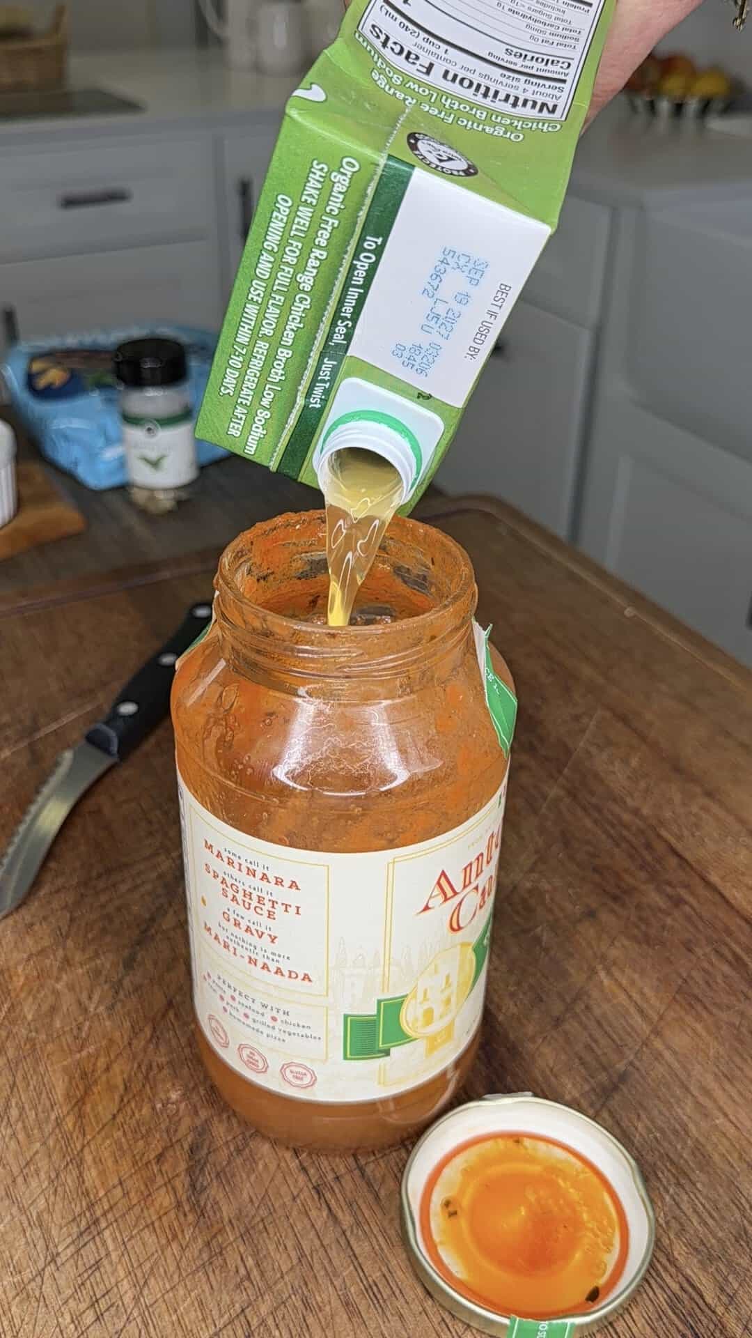 A hand pours liquid from a green carton into an open jar of pasta sauce on a wooden counter. The jar lid is beside it and a knife is nearby. Kitchen items are visible in the background.