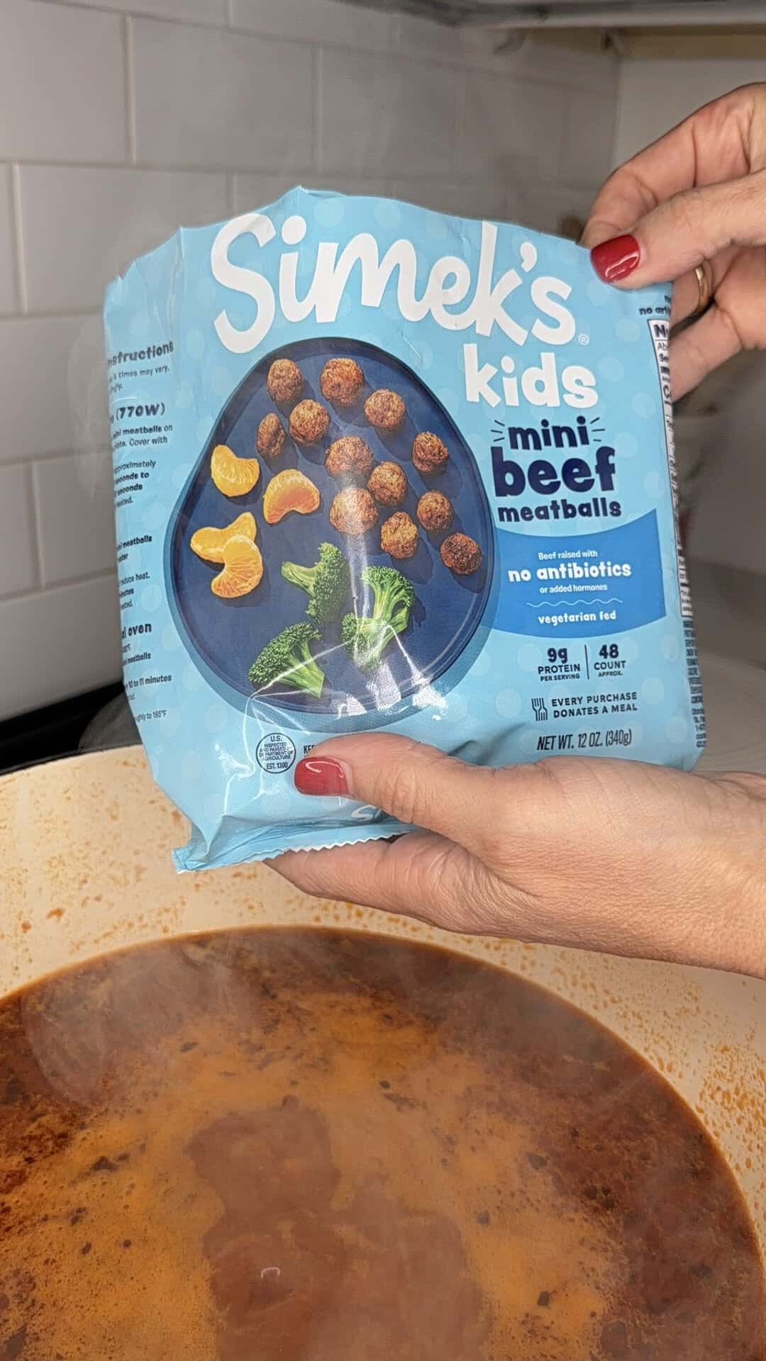A person with red nails holds a blue bag of Simeks Kids Mini Beef Meatballs over a pot of soup or sauce on the stove. The bag features images of meatballs and vegetables.