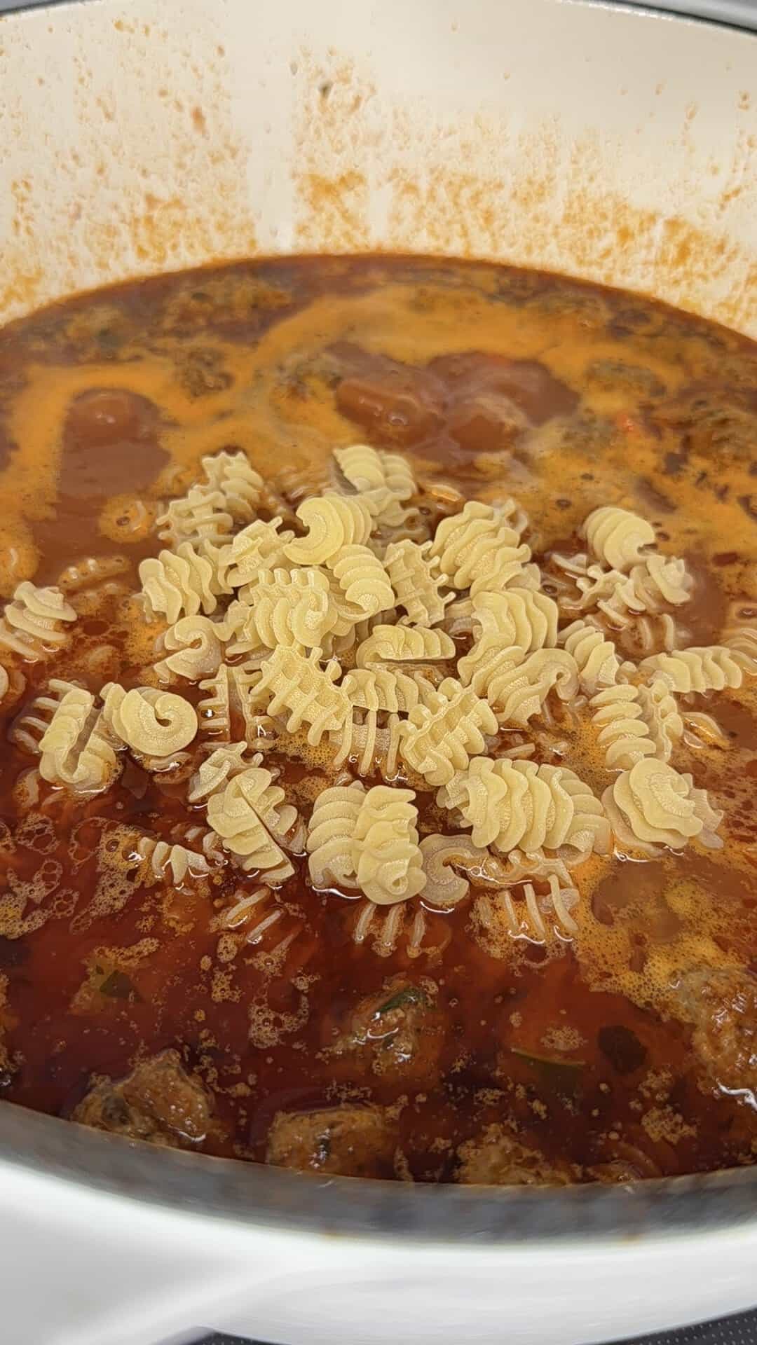A pot of tomato-based soup with dry pasta in fun shapes, including hashtags and at symbols, scattered on top before being stirred in.