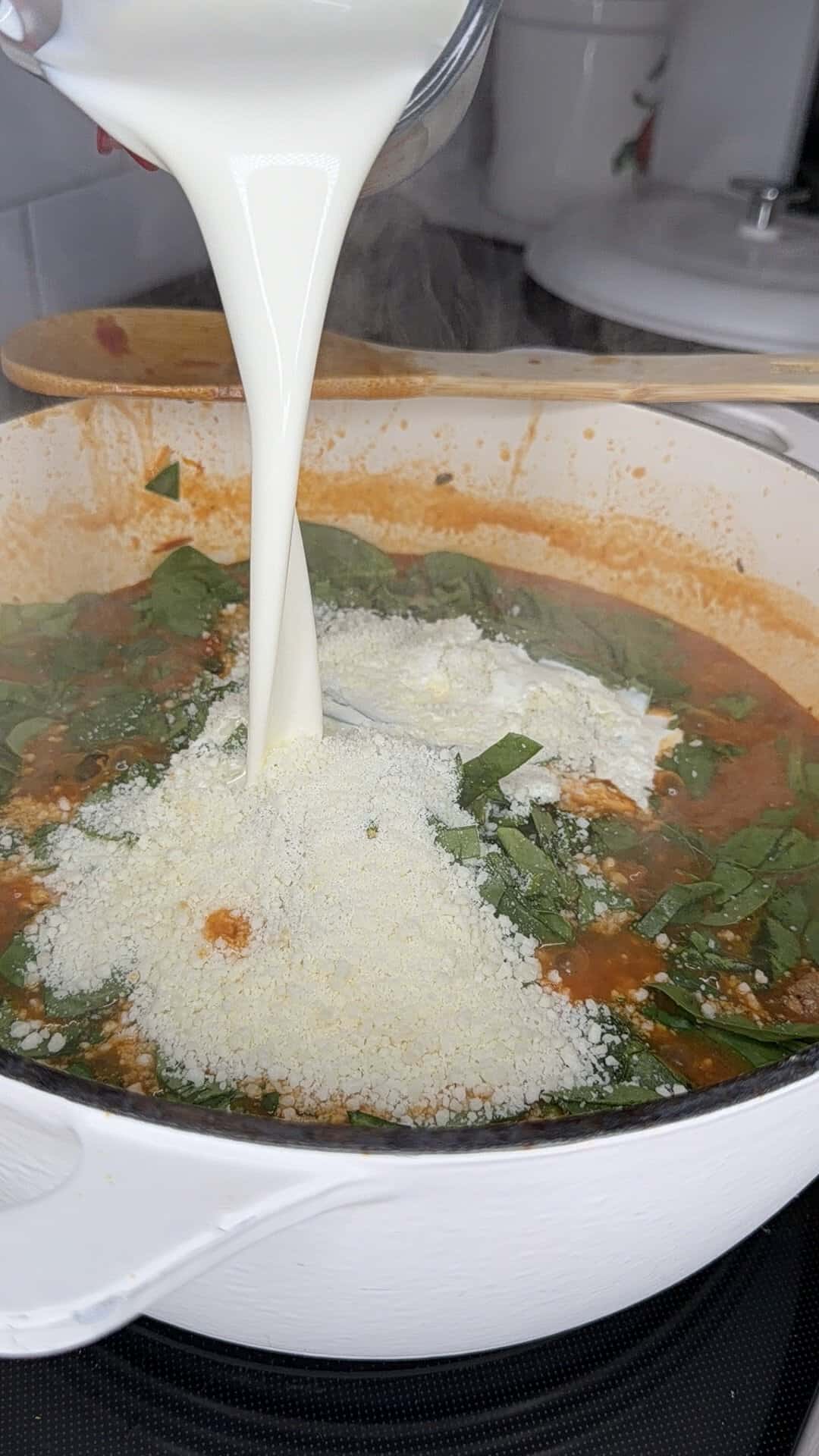 Milk is being poured from a glass measuring cup into a white pot containing tomato sauce, spinach, and grated cheese on a stovetop. A wooden spoon and kitchen items are visible in the background.