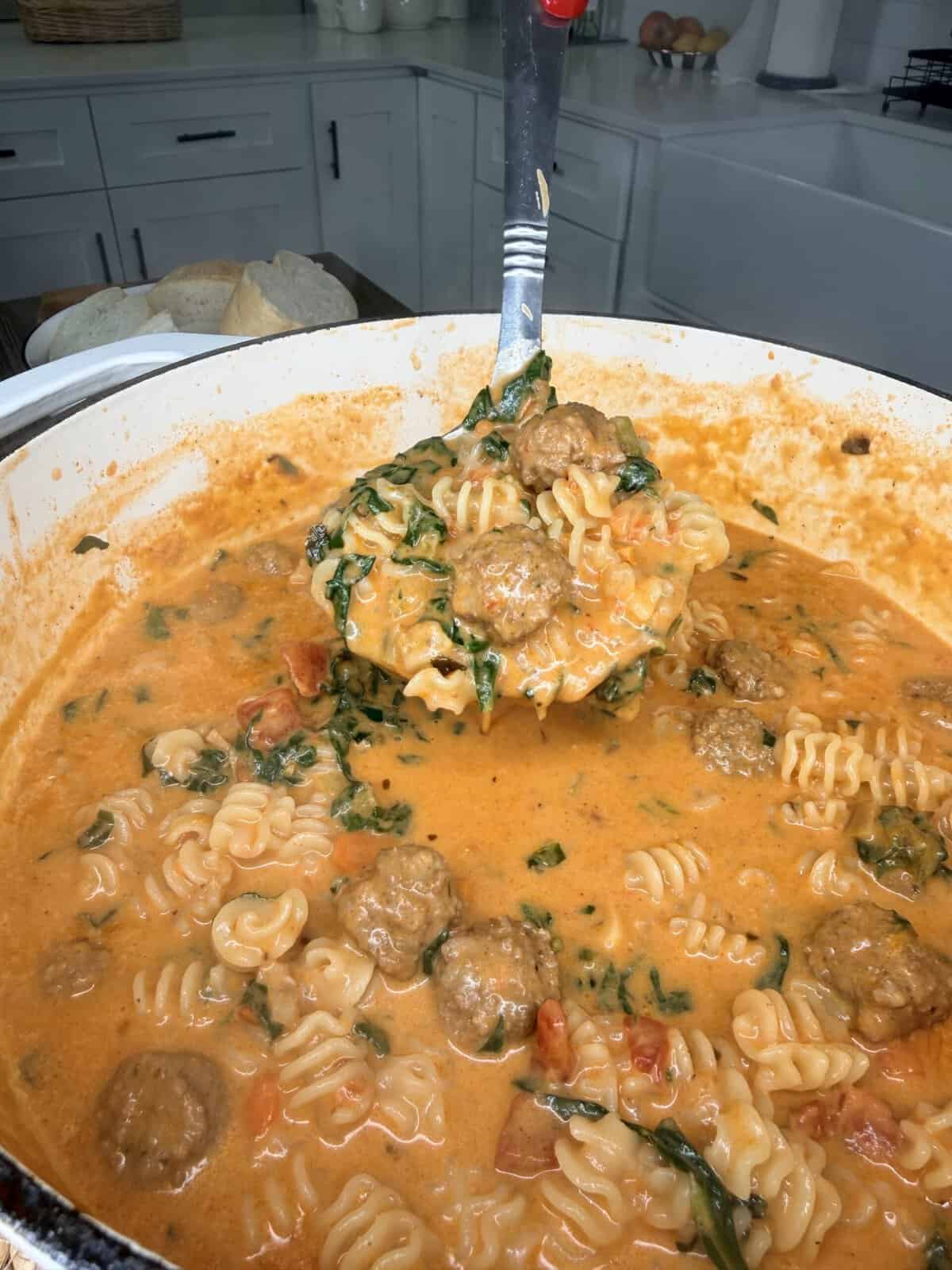 A close-up of a pot filled with creamy tomato pasta, meatballs, spinach, and ruffled noodles. A ladle lifts a serving above the pot. Bread is visible in the background on a kitchen counter.