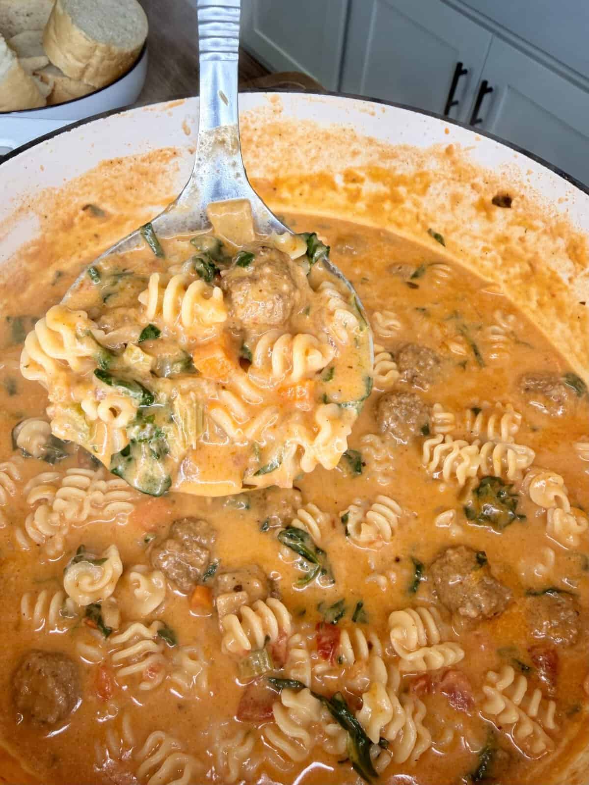 A ladle scoops creamy, tomato-based pasta with radiatori noodles, meatballs, and greens from a large pot. Sliced bread sits on a plate nearby.