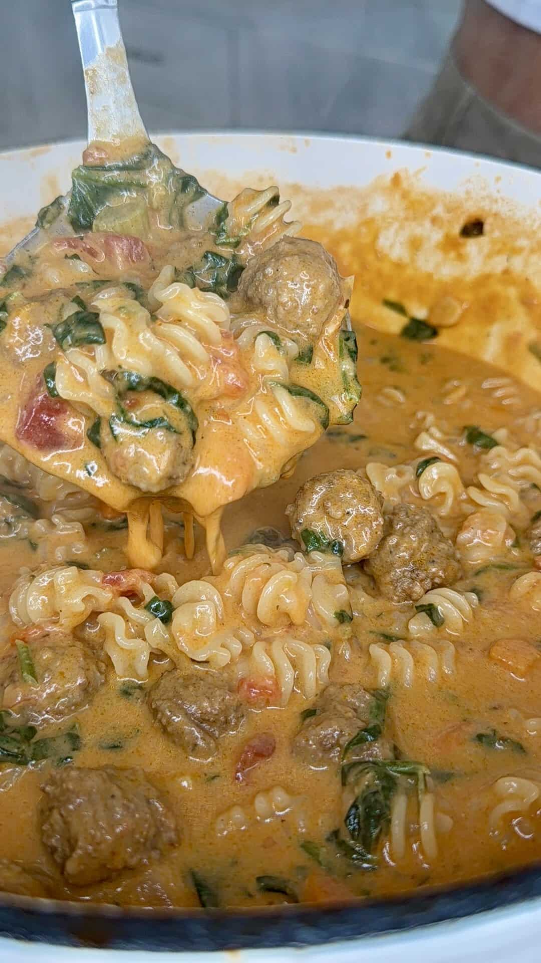 A close-up of creamy soup with curly noodles, meatballs, chopped greens, and tomatoes, being scooped with a ladle from a large white pot.
