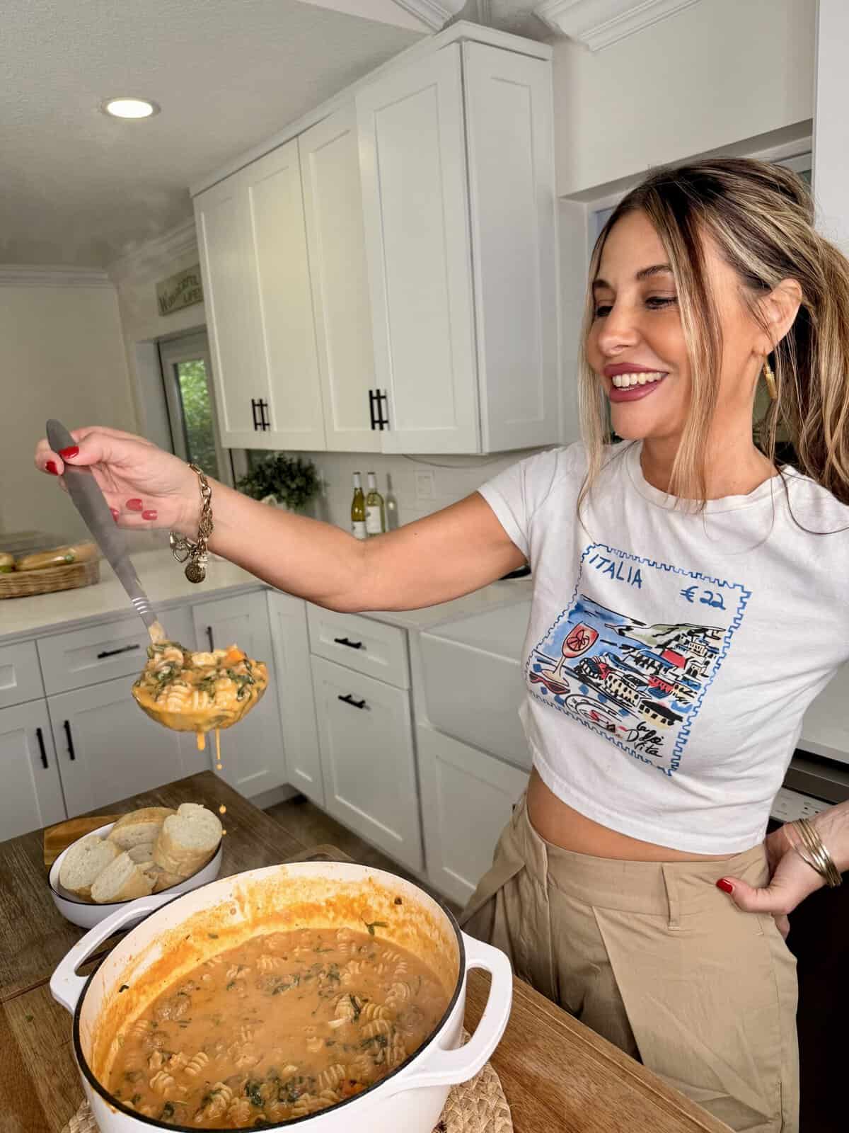 A woman smiling in a kitchen ladles soup from a white pot on the counter. Sliced bread is arranged on a plate beside the pot. White cabinets and kitchen decor are visible in the background.