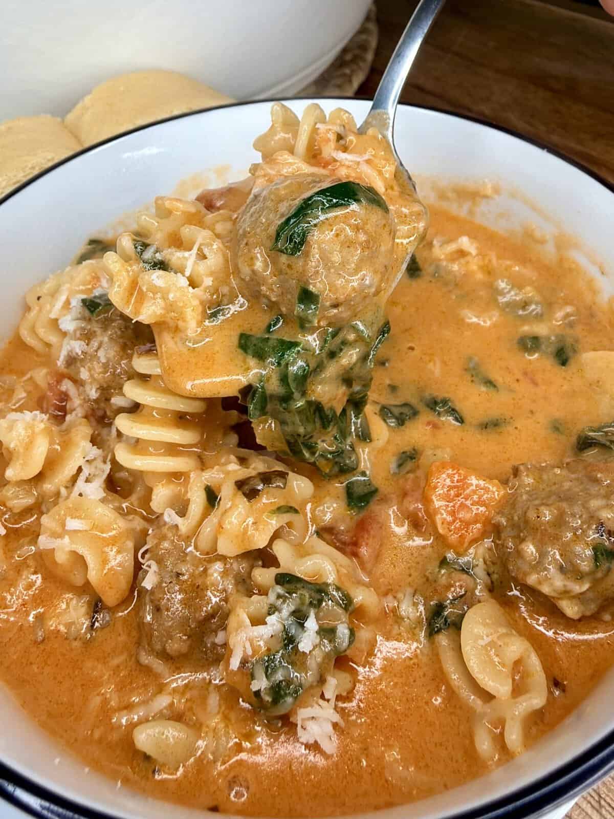 A close-up of a bowl of creamy tomato soup with pasta, spinach, meatballs, and grated cheese. A spoon is lifting a portion of soup from the bowl.