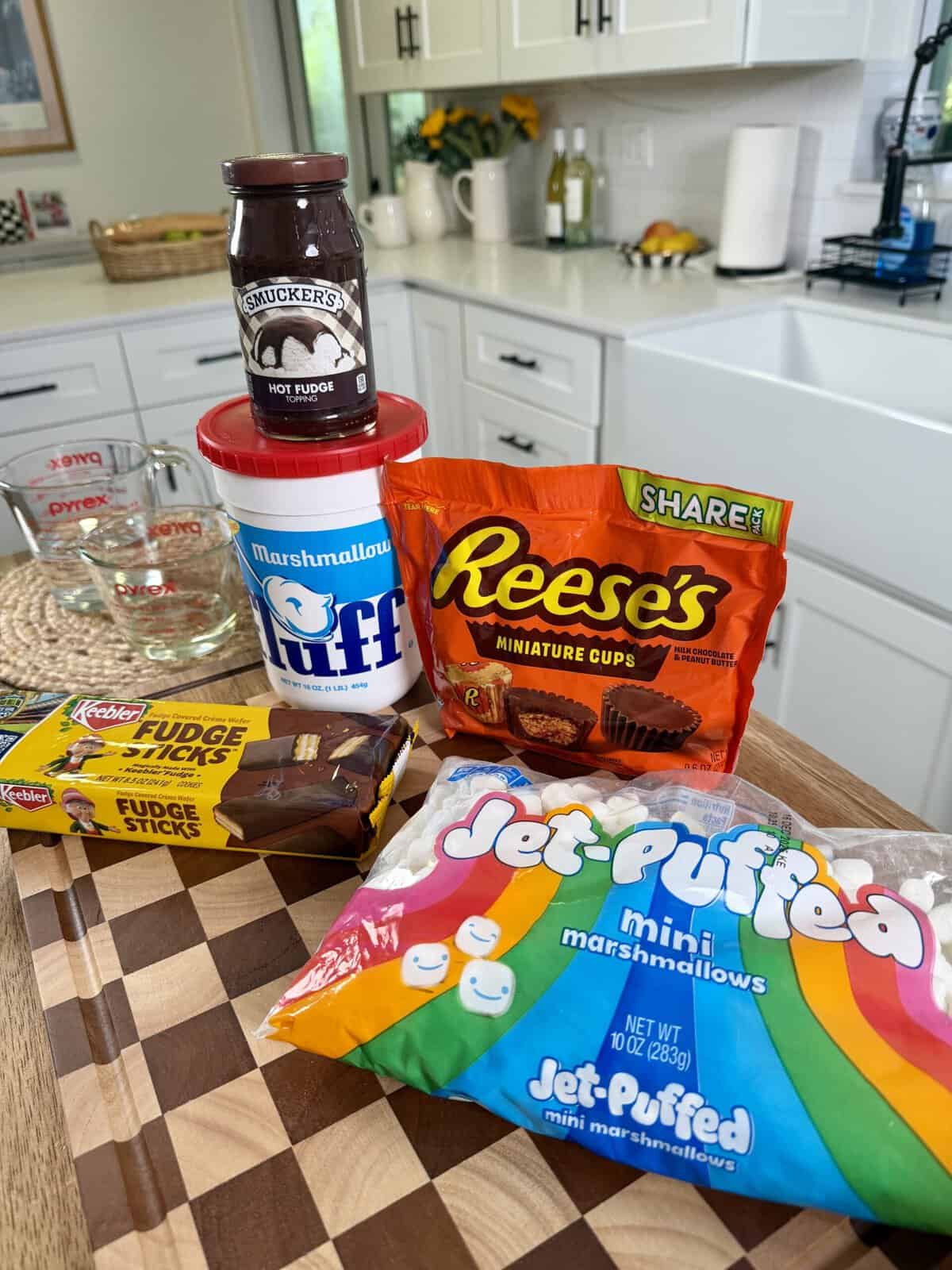 Assorted dessert ingredients on a kitchen counter, including marshmallow fluff, mini Reeses cups, fudge sticks, hot fudge, and mini marshmallows, with measuring cups and a cutting board nearby.
