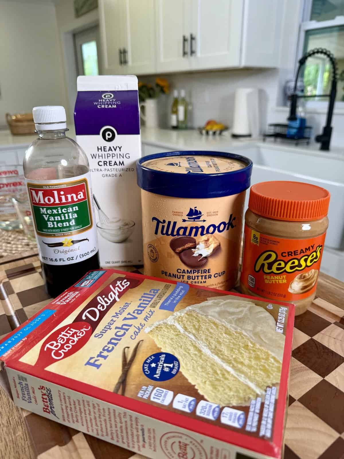 Five dessert ingredients—vanilla extract, heavy whipping cream, Tillamook ice cream, Reeses creamy peanut butter, and Betty Crocker French vanilla cake mix—are arranged on a kitchen counter in front of white cabinets and a sink.