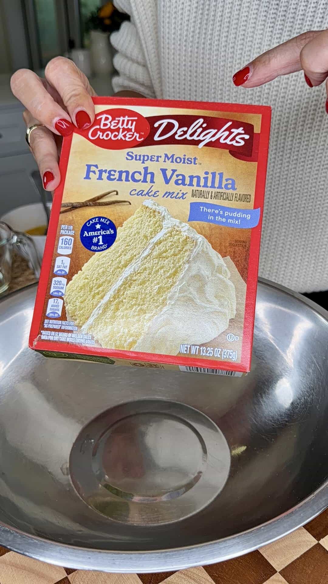 A person with red-painted nails holds a box of Betty Crocker Delights Super Moist French Vanilla cake mix above a large empty metal bowl.