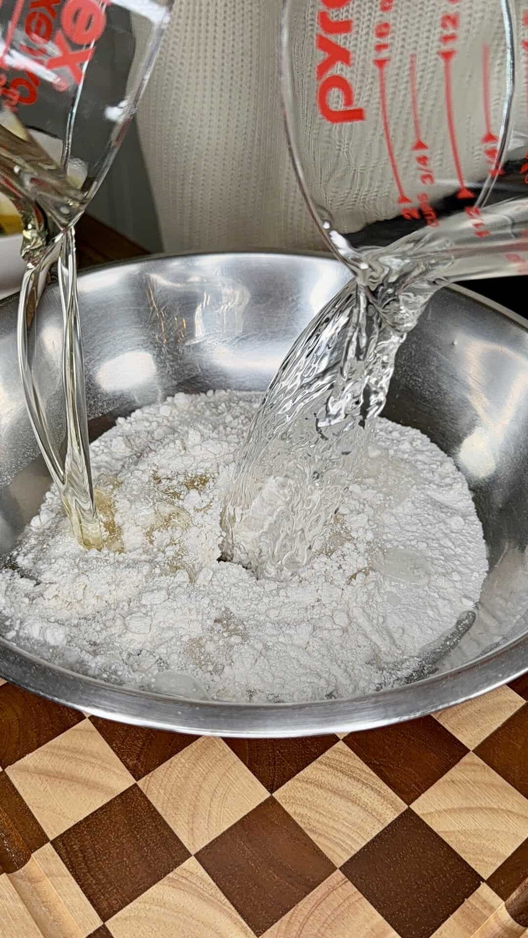 A close-up of two measuring cups pouring liquid into a metal bowl filled with flour, set on a checkered wooden surface.