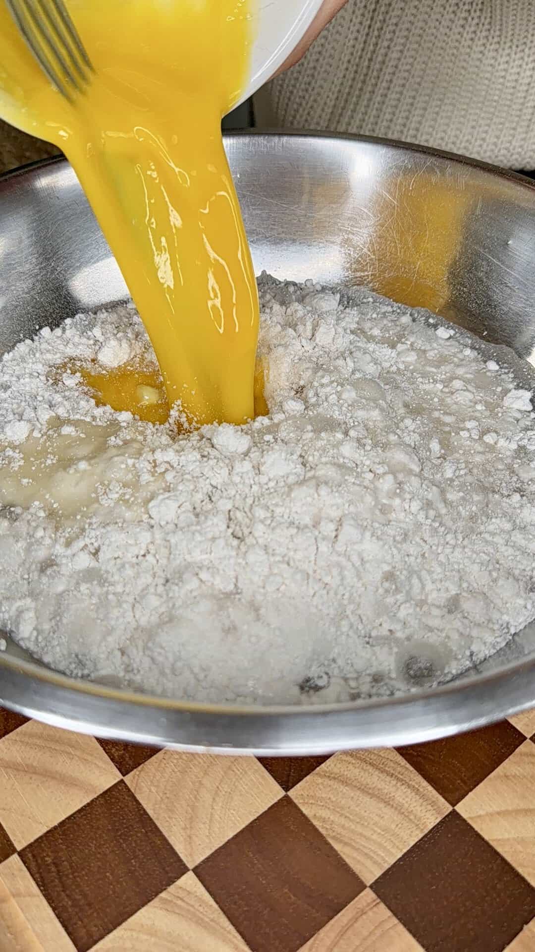 A person pours beaten eggs into a metal bowl filled with flour on a wooden checkered surface, preparing to mix baking ingredients.