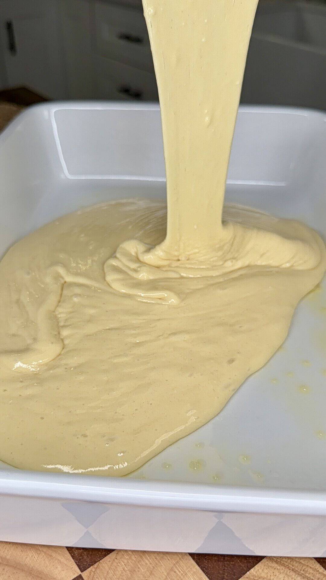 Thick yellow batter being poured into a white baking dish on a wooden surface, ready for baking.