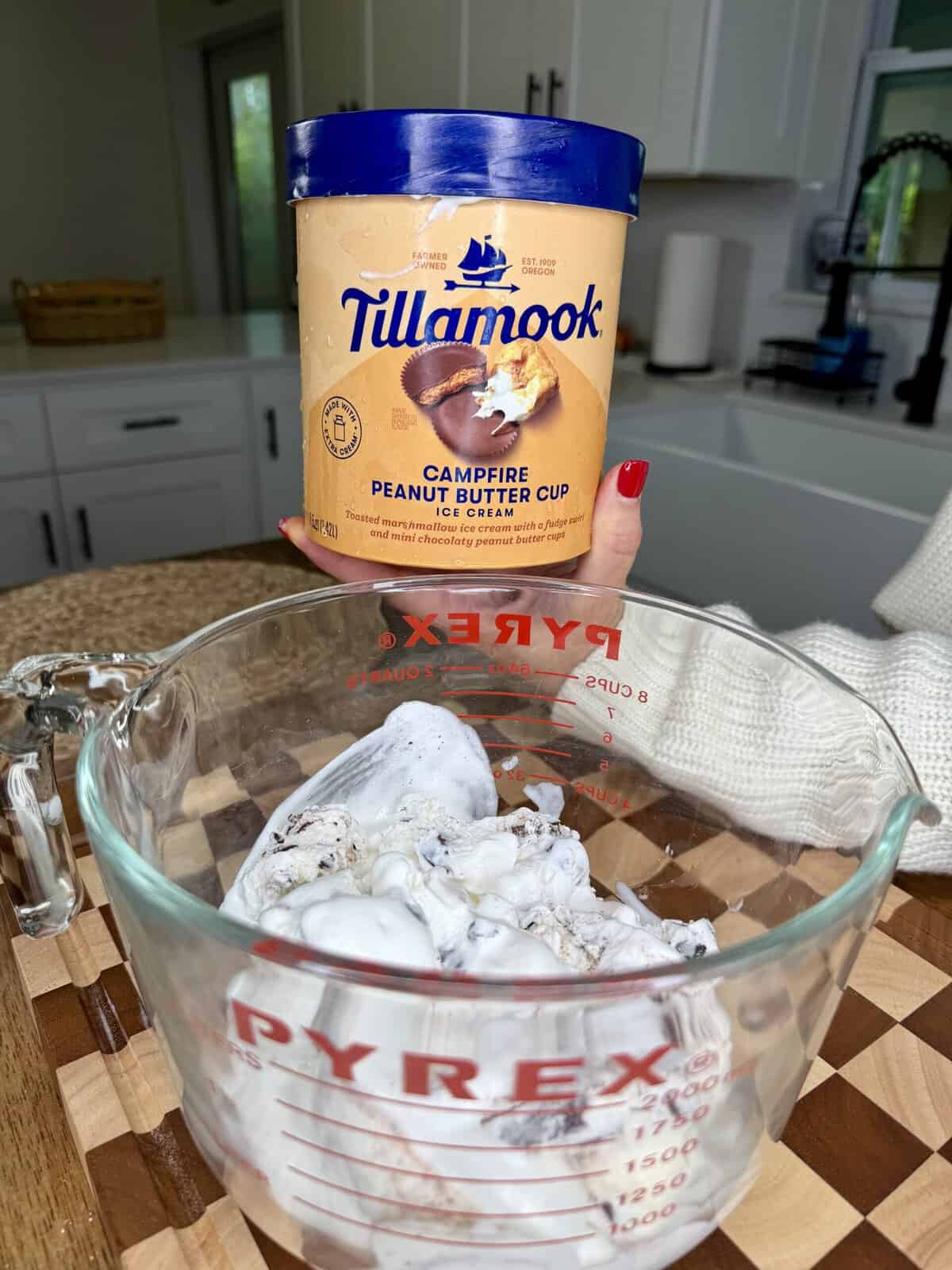 A person holds a Tillamook Campfire Peanut Butter Cup ice cream container above a Pyrex measuring cup filled with a similar ice cream, in a bright kitchen setting.