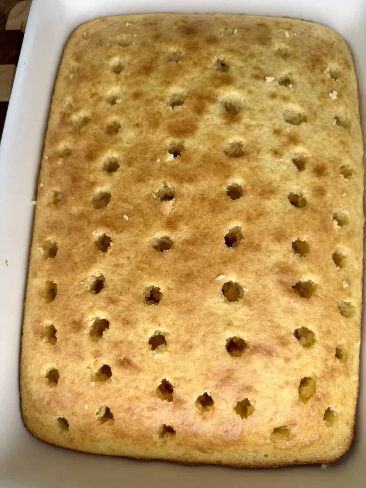 A golden-brown sheet cake in a white baking dish, with evenly spaced holes poked across its surface, ready for adding filling or topping.