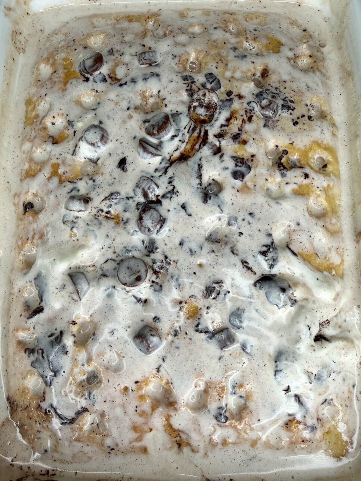 A close-up of a baked dessert with a melted, creamy marshmallow topping, visible chocolate pieces, and yellowish cake peeking through underneath. The surface looks gooey, swirled, and lightly toasted in spots.
