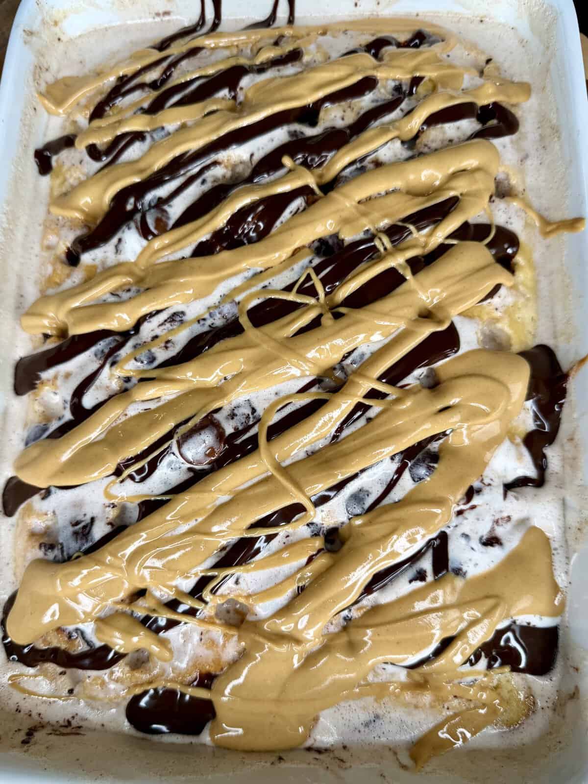 A close-up of a dessert topped with thick drizzles of chocolate and peanut butter sauce, spread over a creamy or frosted surface in a rectangular dish.