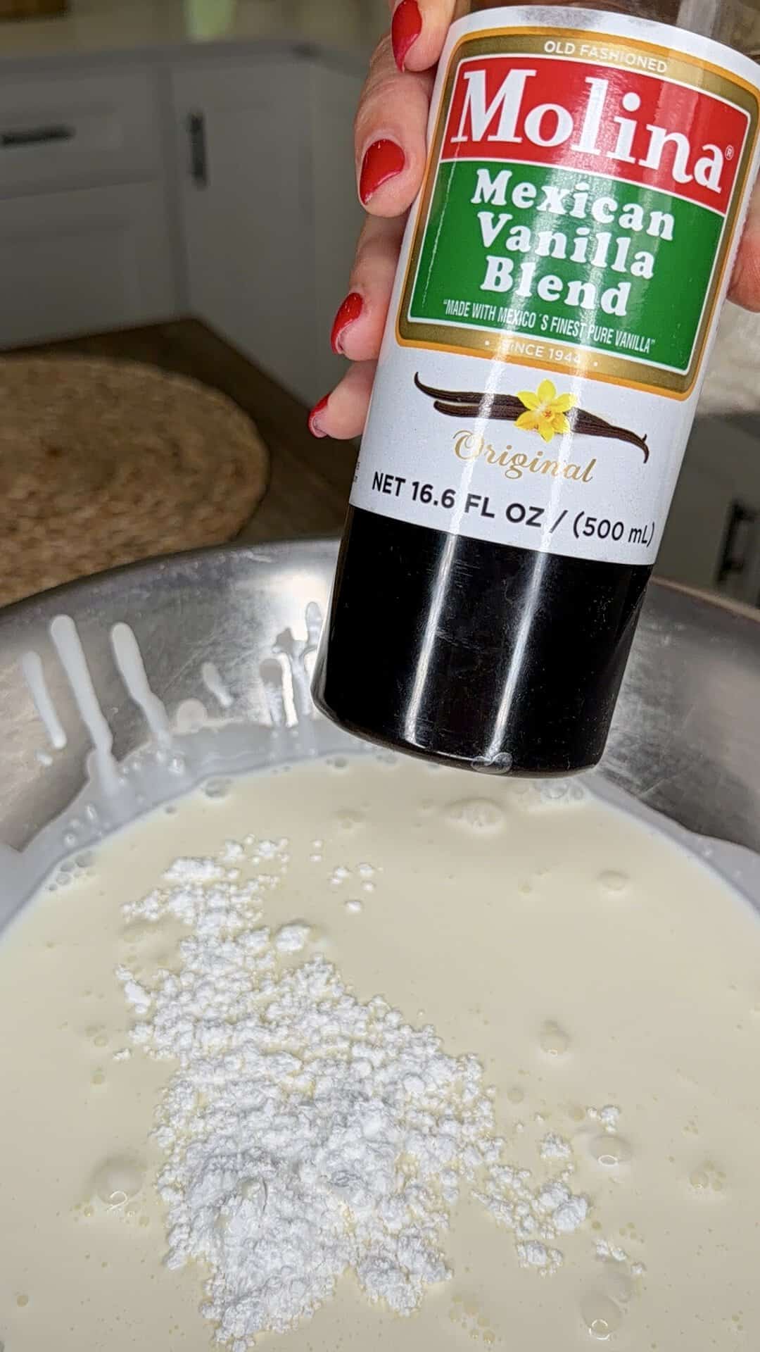 A hand holds a bottle of Molina Mexican Vanilla Blend over a metal bowl filled with cream and powdered sugar, ready for mixing.