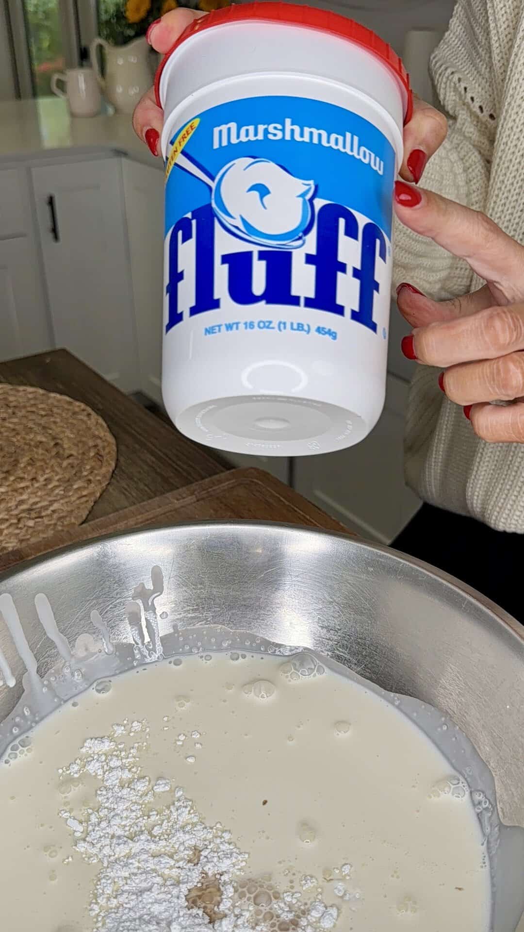 A person holds a container of Marshmallow Fluff above a mixing bowl filled with powdered sugar, milk, and other ingredients on a kitchen counter.