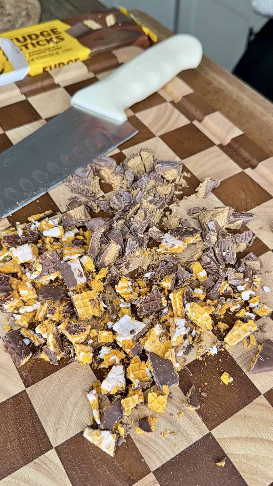Chopped chocolate candy pieces with fudge and honeycomb on a checkered wooden cutting board, next to a white-handled knife.