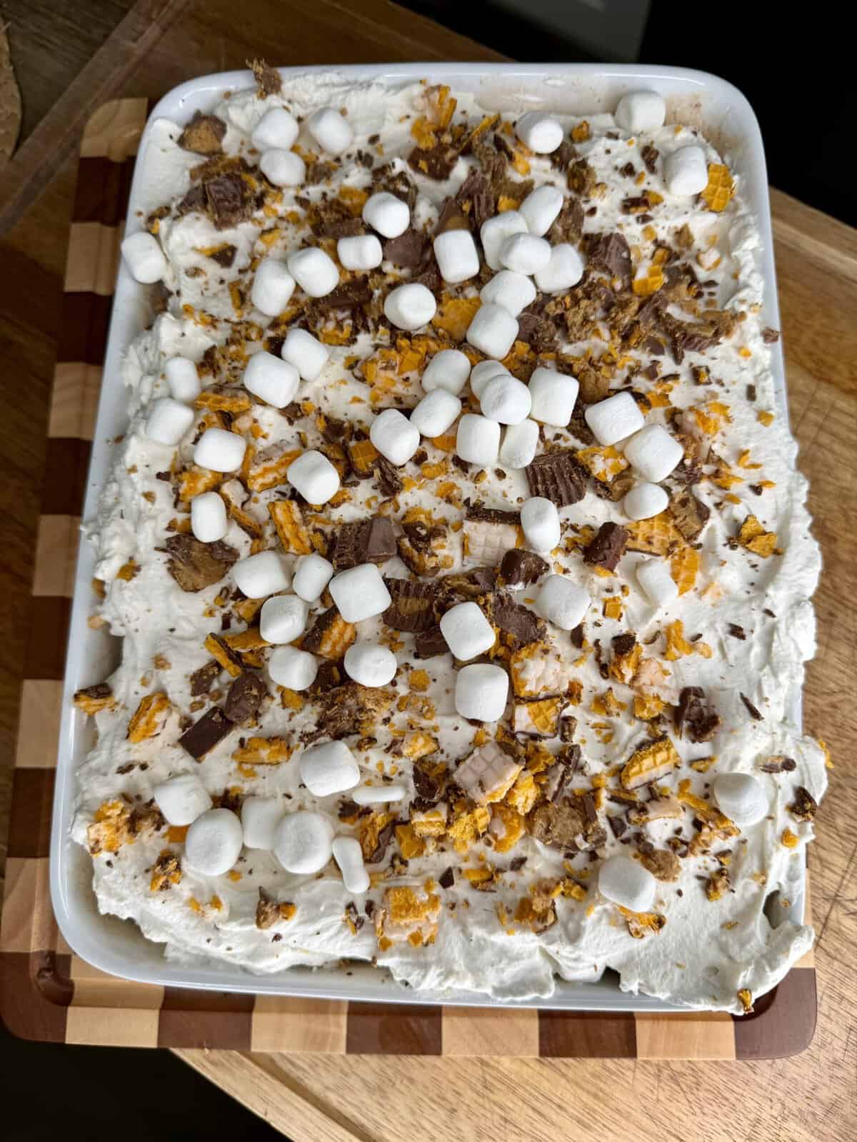 A rectangular dessert with a whipped topping, mini marshmallows, and crumbled chocolate and orange-colored pieces, served on a wooden cutting board.