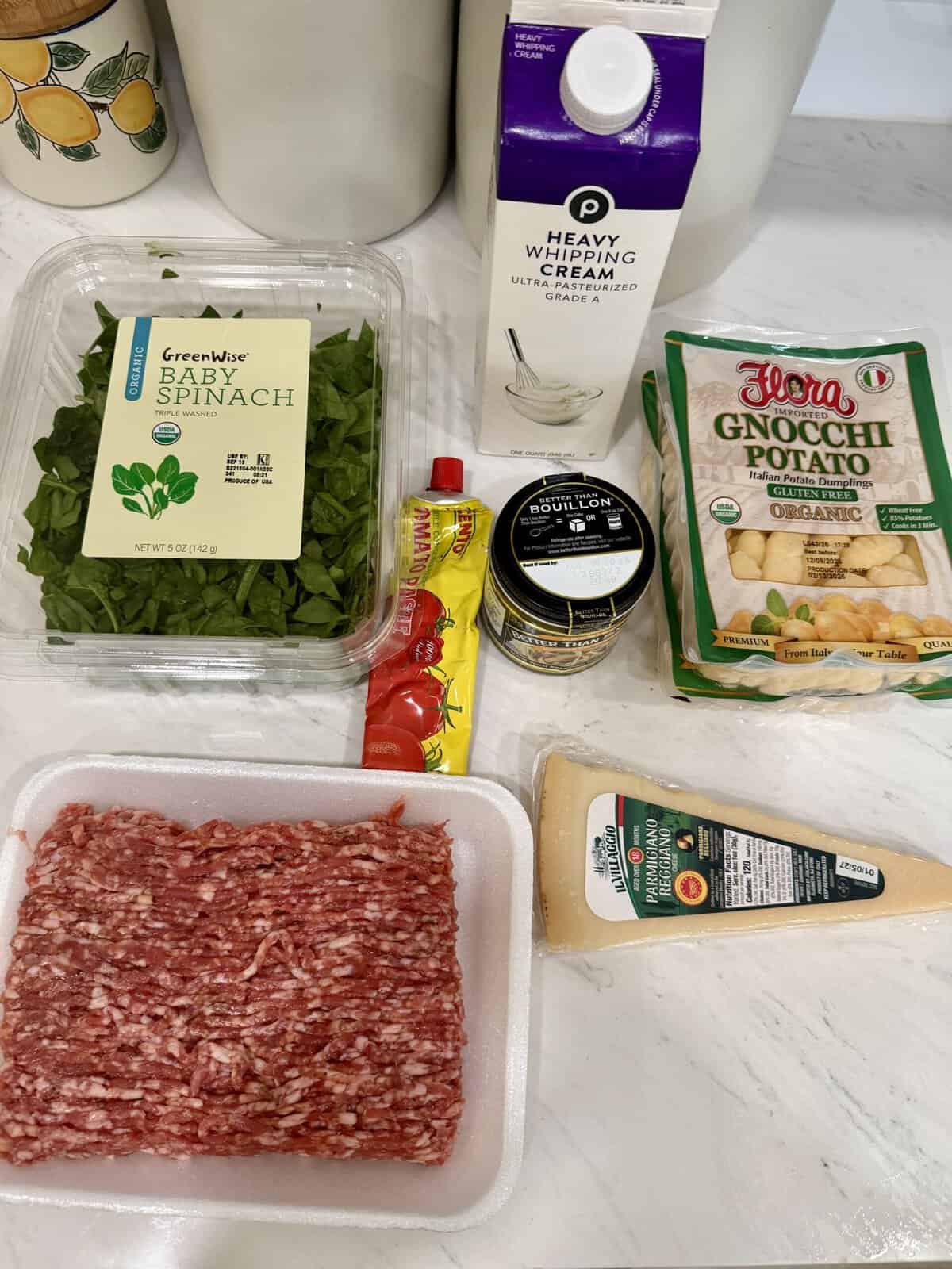 Various ingredients on a kitchen counter: ground meat, baby spinach, heavy cream, potato gnocchi, Parmesan cheese, tomato paste, and garlic bouillon paste.