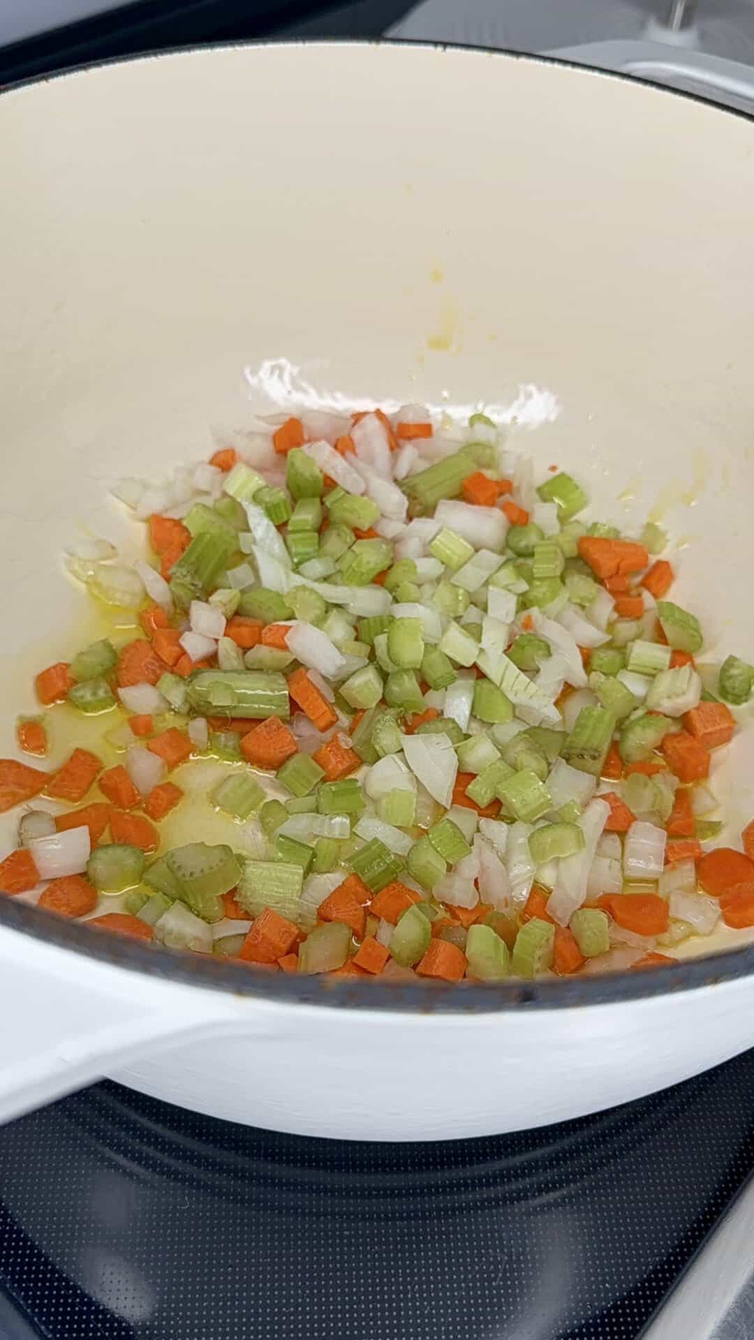 Diced carrots, celery, and onions are being sautéed in a white pot on a stovetop.
