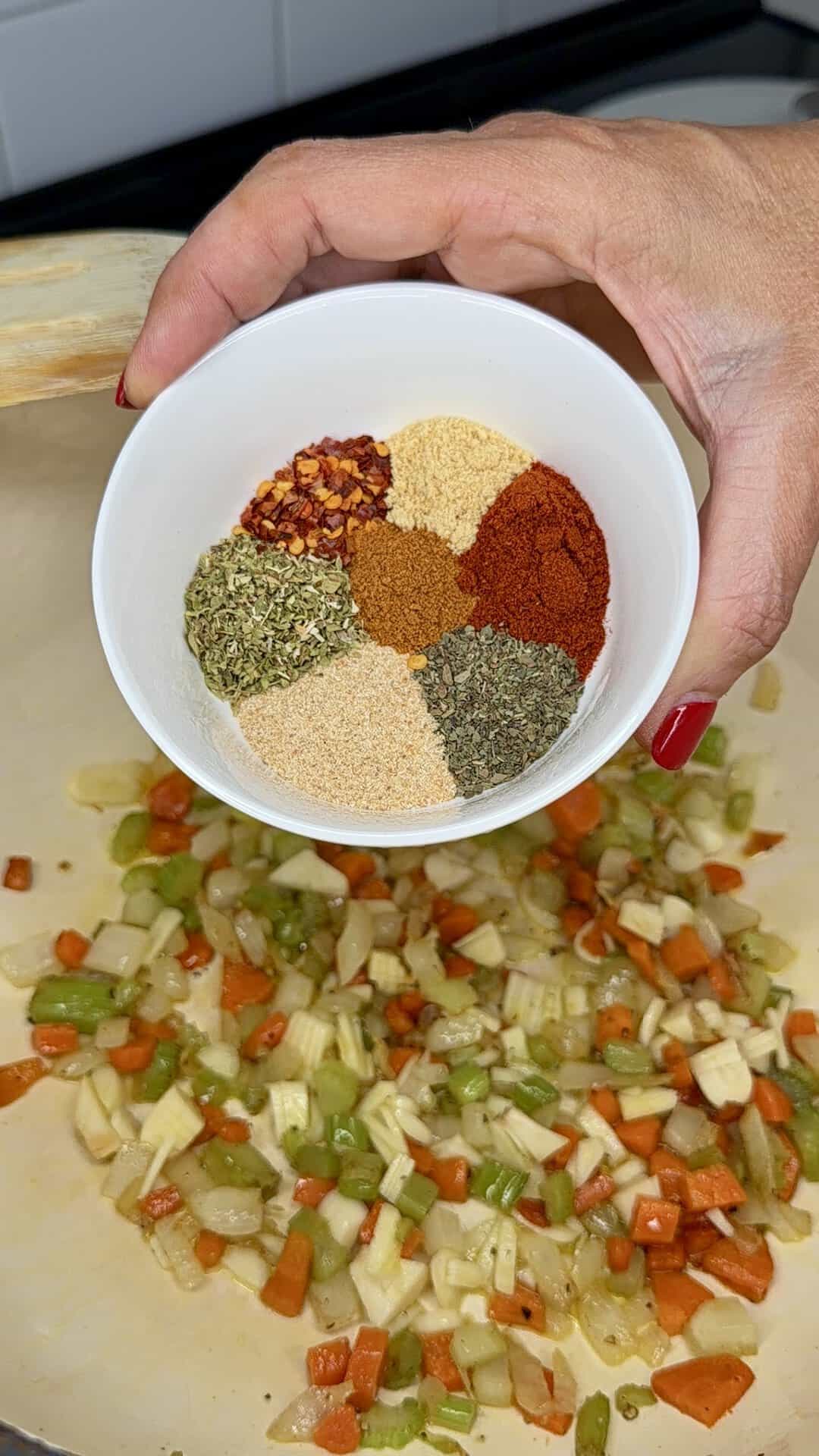 A hand holds a bowl of six assorted spices above a pan of sautéed chopped onions, carrots, green peppers, and garlic.