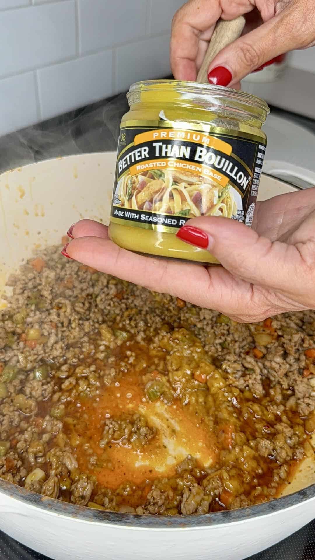 A person holds a jar of Better Than Bouillon roasted chicken base over a pot of cooked ground meat and vegetables, stirring with a wooden spoon.