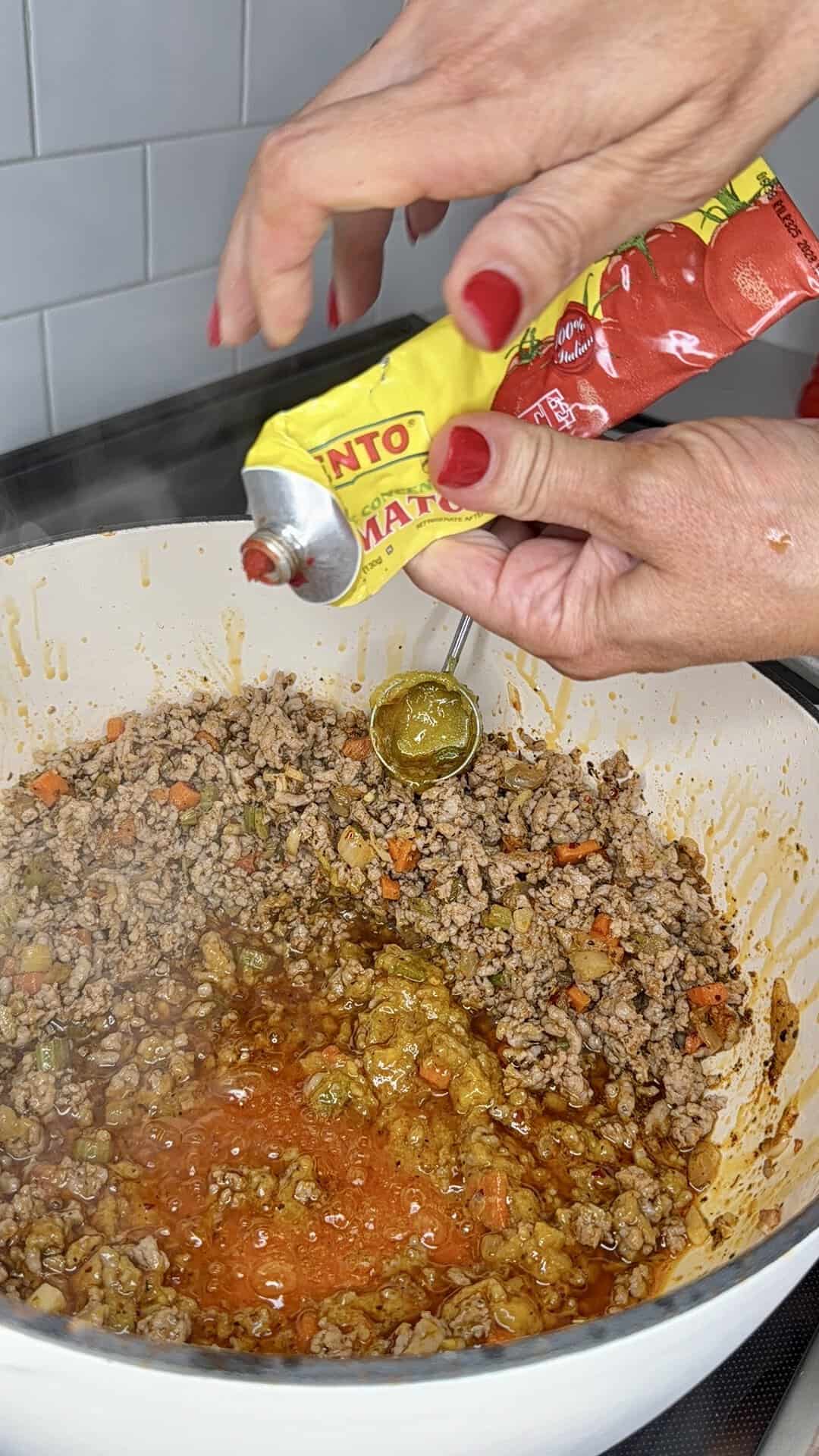 Two hands with red nails squeeze tomato paste from a yellow tube into a white pot filled with browned ground meat, vegetables, and sauce, while a spoon holds some paste over the mixture.