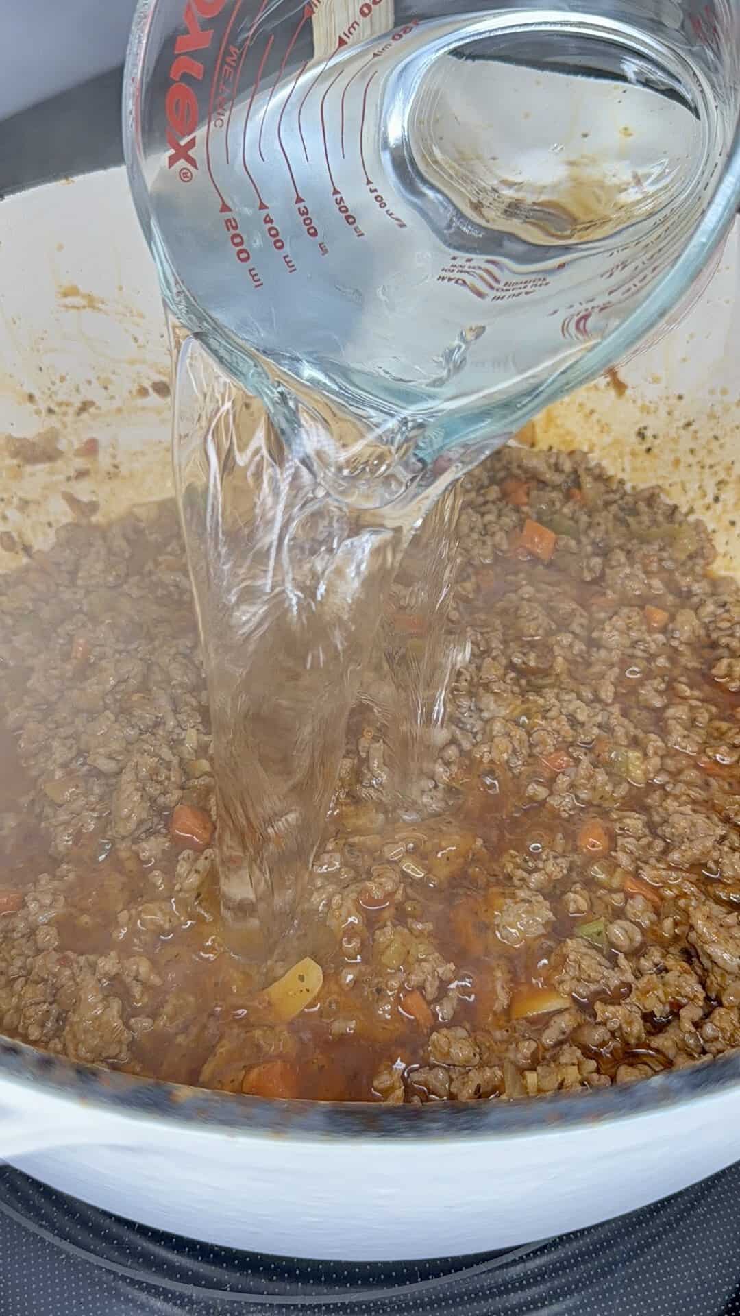 Water being poured from a glass measuring cup into a pot filled with cooked ground meat, carrots, and sauce, as part of a cooking process.