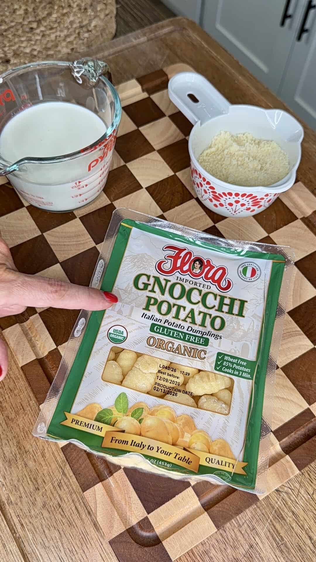A hand points to a package of Flora Organic Gnocchi Potato on a wooden cutting board, next to a measuring cup of milk and a small bowl of grated cheese.