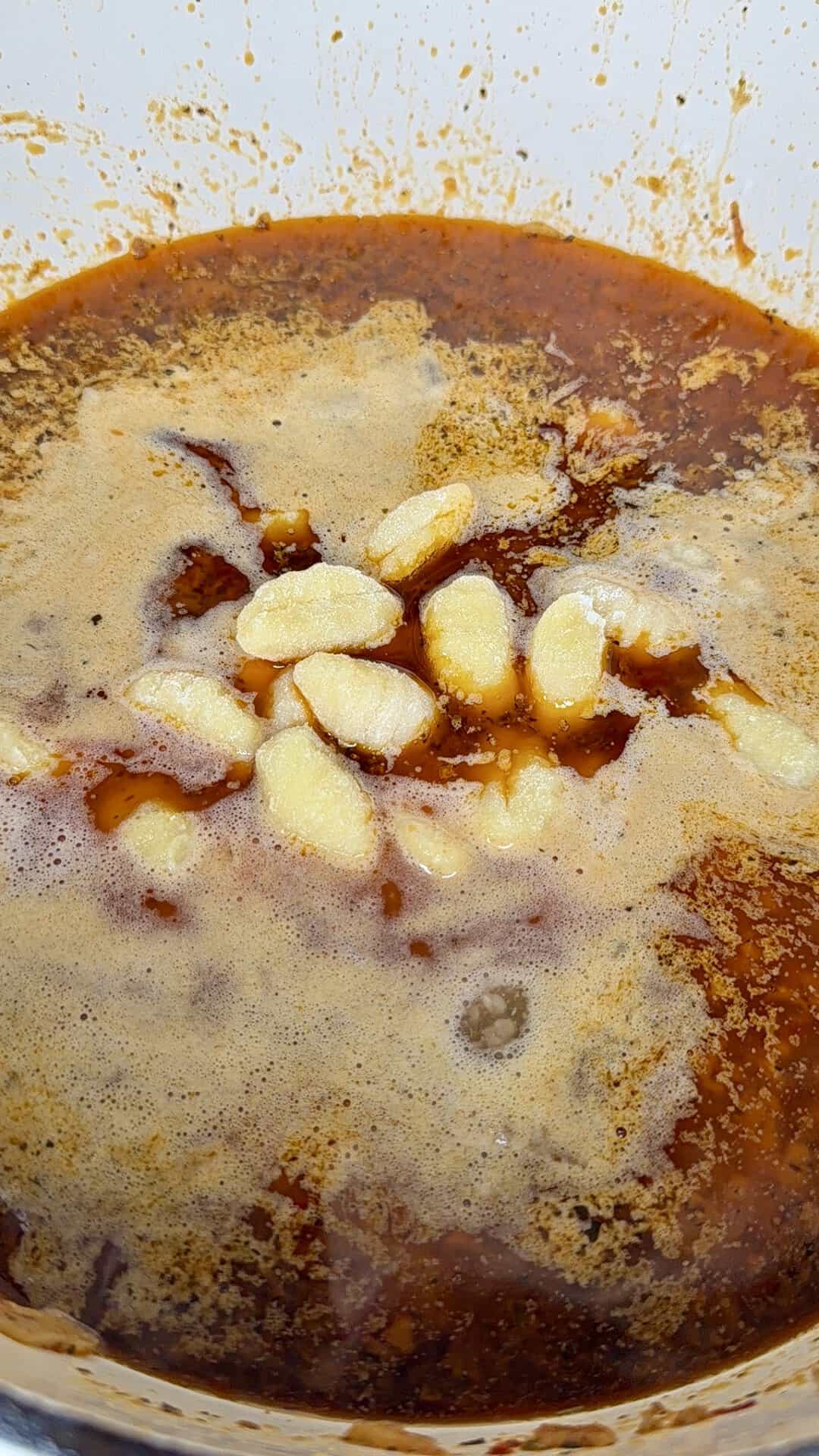 A close-up of a pot filled with red stew, topped with bubbling oil and several whole peeled garlic cloves floating on the surface.