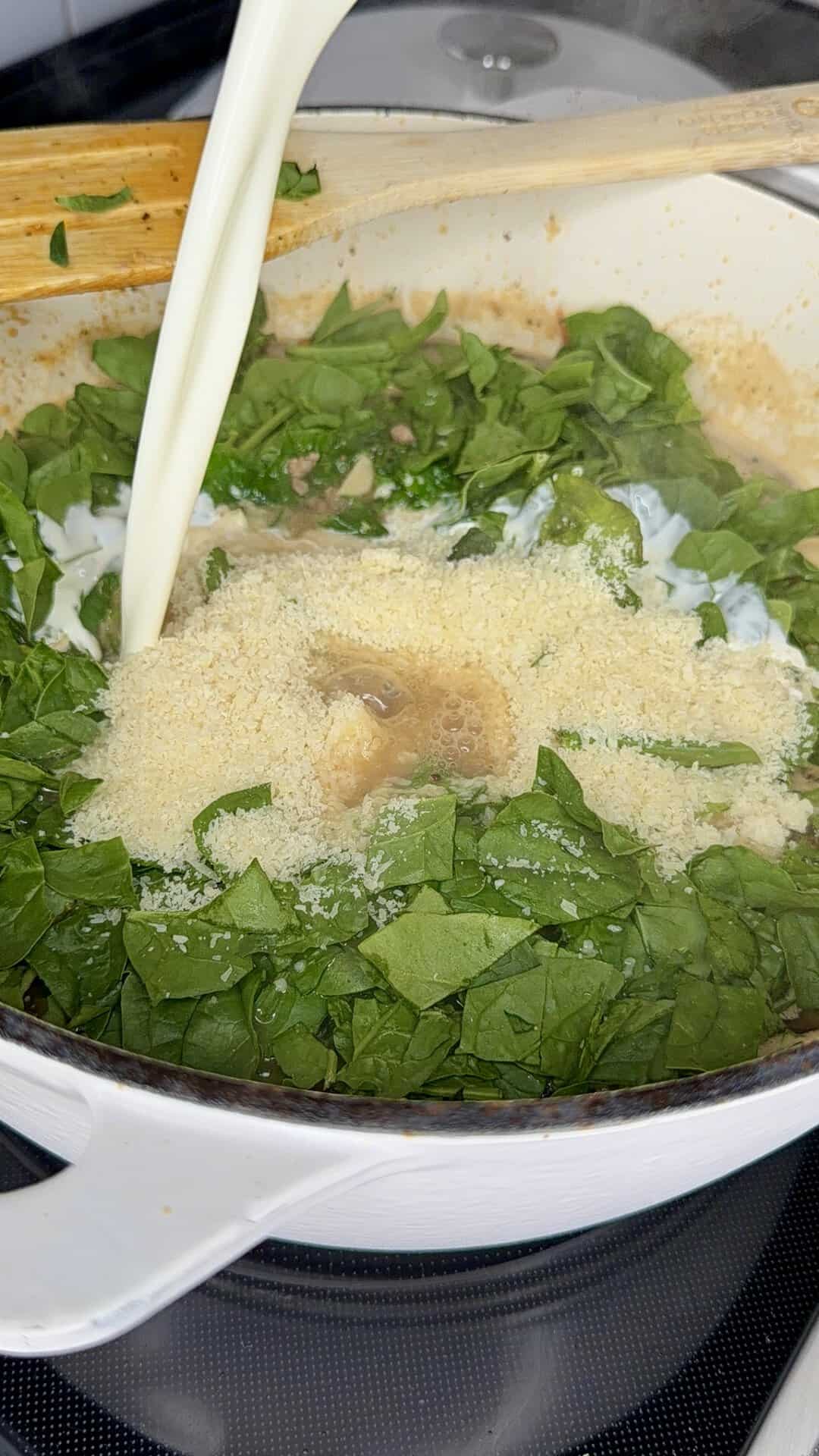 Milk is being poured into a white pot containing chopped spinach, grated cheese, and other ingredients. A wooden spoon rests on the side of the pot, and the mixture appears to be mid-cooking.