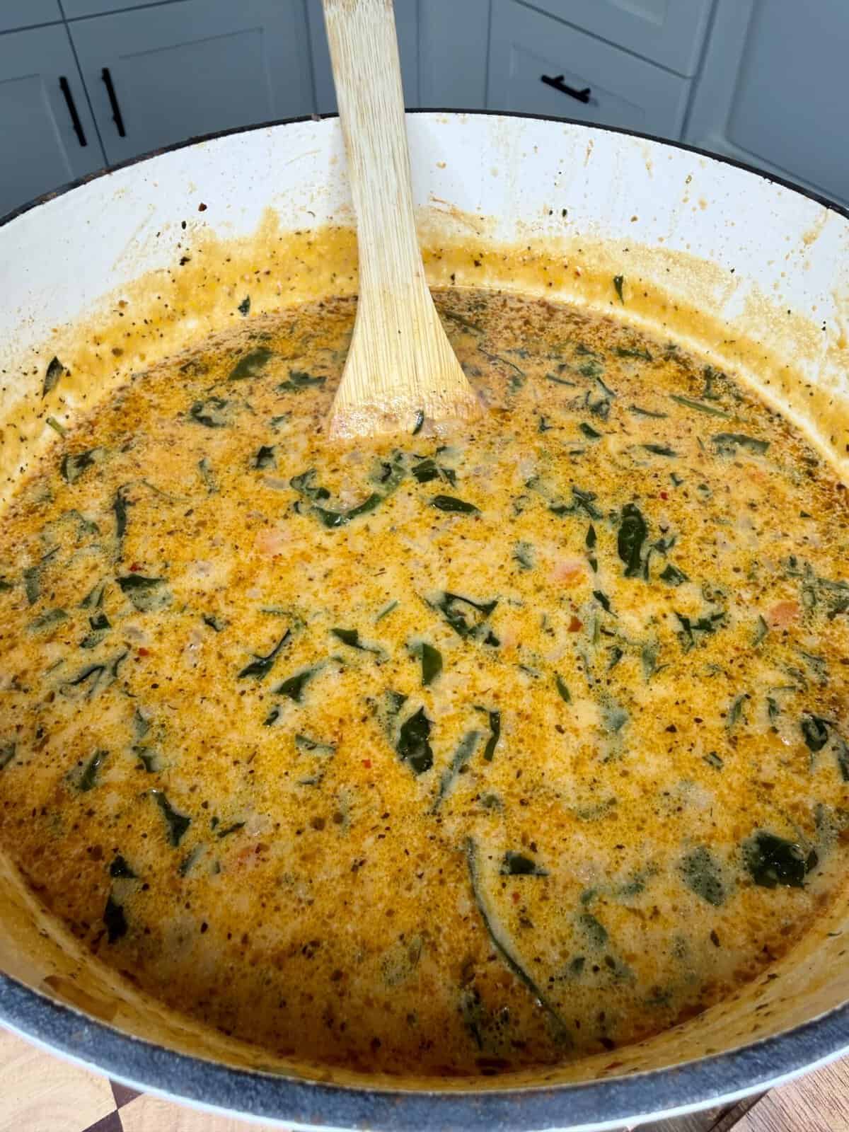 A large pot filled with creamy orange soup containing green leafy vegetables and small chunks, being stirred with a wooden spoon in a kitchen setting.