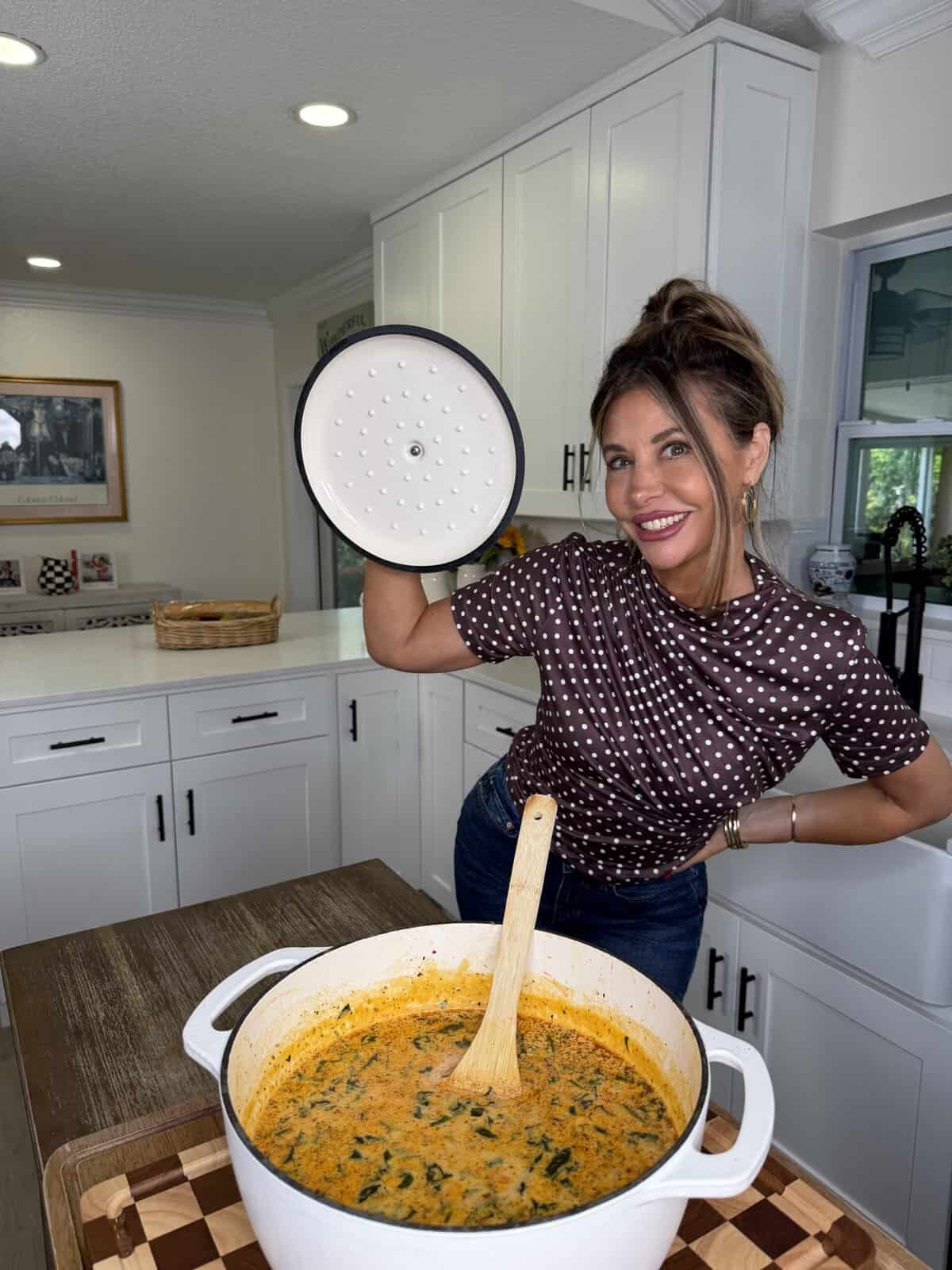 A woman in a polka dot shirt smiles while holding a pot lid above a white pot filled with soup or stew in a bright, modern kitchen. A wooden spoon rests in the pot on a wooden table.