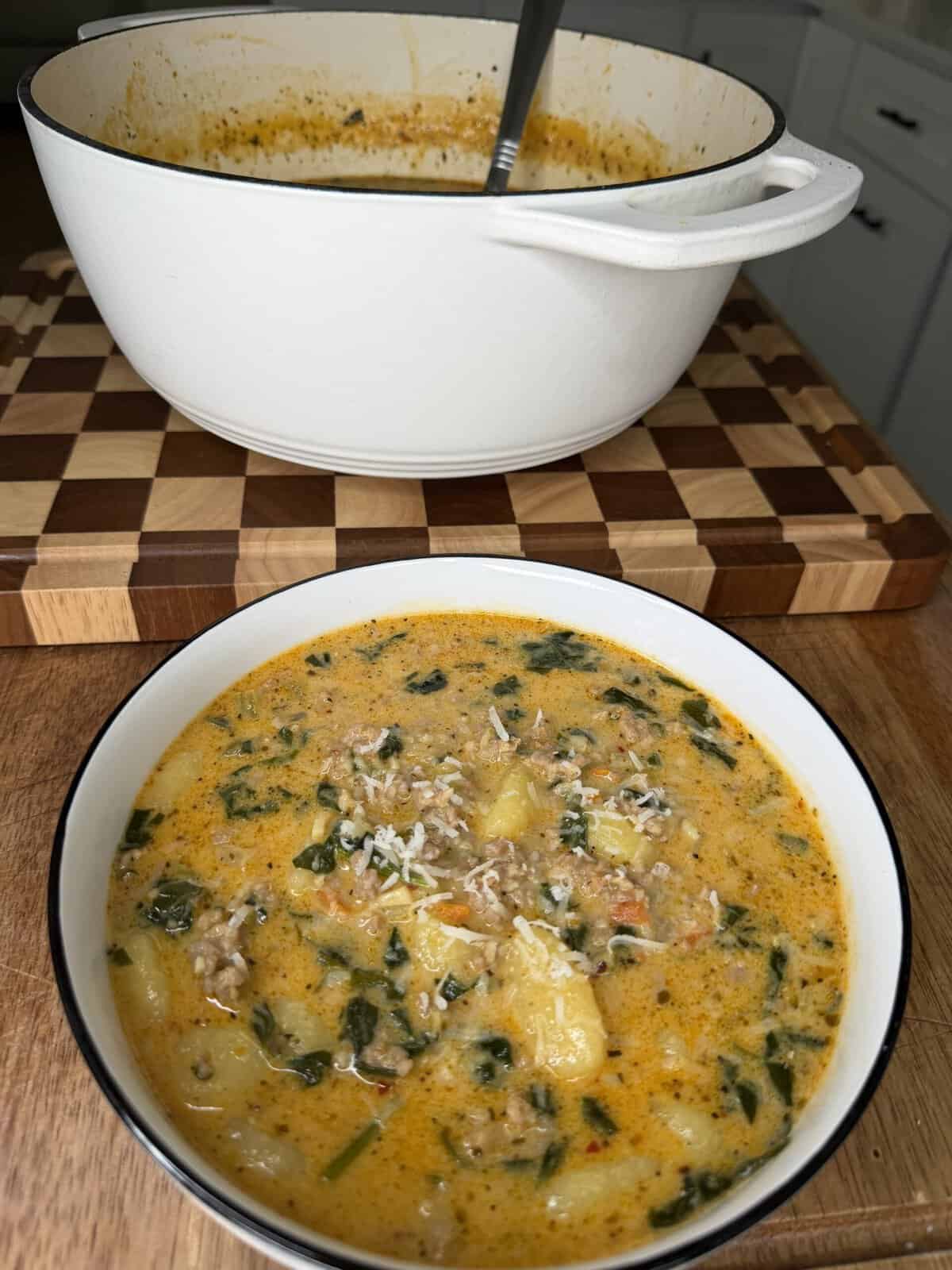 A bowl of creamy soup with sausage, potatoes, and greens sits in front of a white pot on a wooden board.