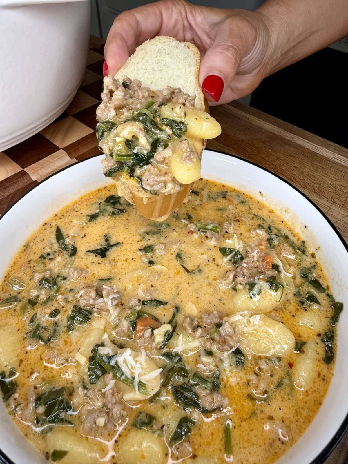 A hand with red nails dips a slice of bread into a bowl of creamy soup with sausage, gnocchi, spinach, and grated cheese on top.
