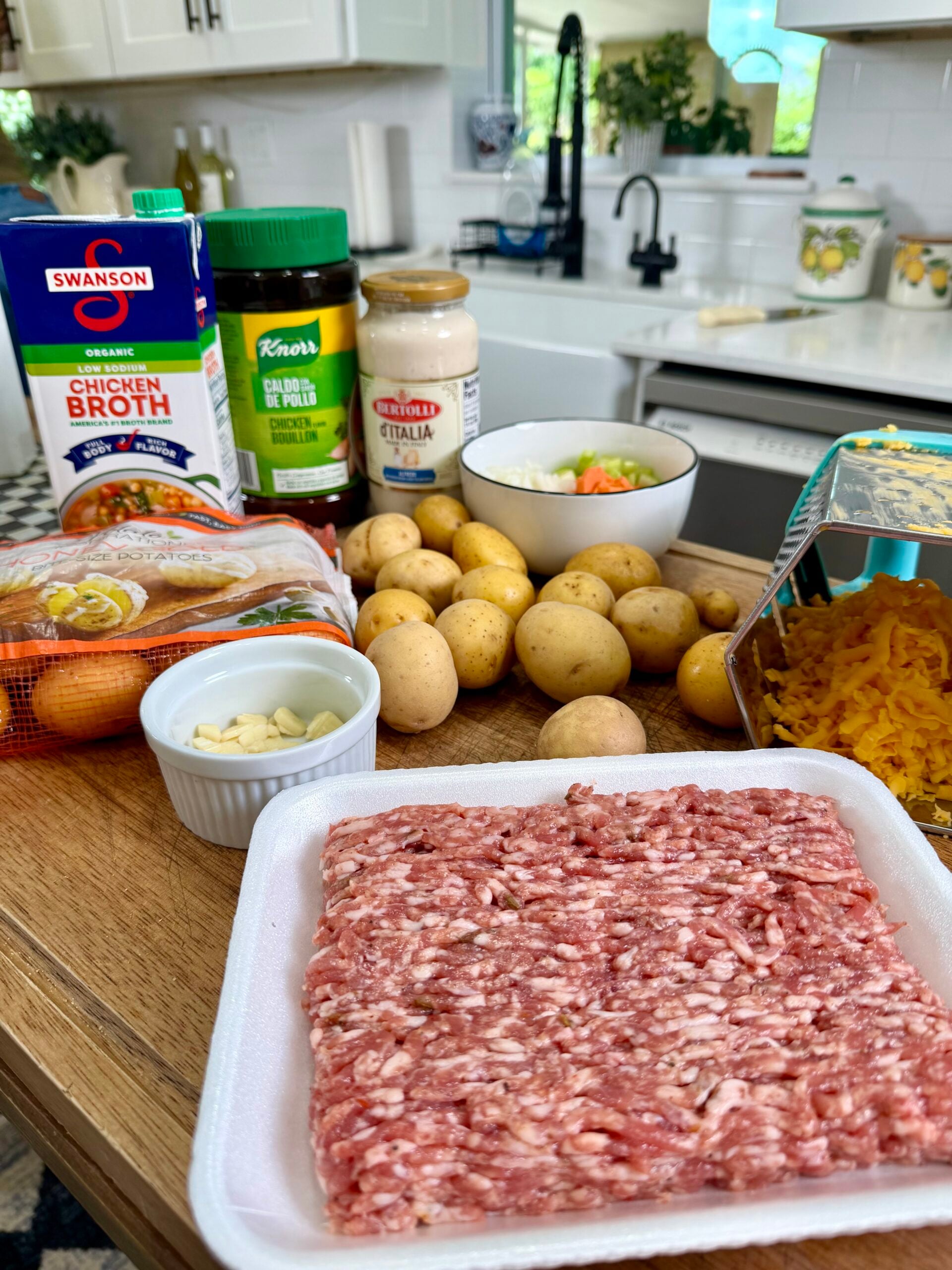 Raw ground pork, baby potatoes, shredded cheddar, garlic, eggs, carrots, celery, broth, Knorr seasoning, and Alfredo sauce are arranged on a white kitchen counter with cabinets and a sink in the background.