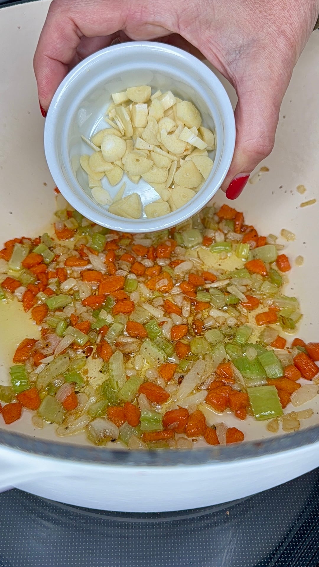 A hand holds a small bowl of sliced garlic above a pot with sautéed carrots, onions, and celery.