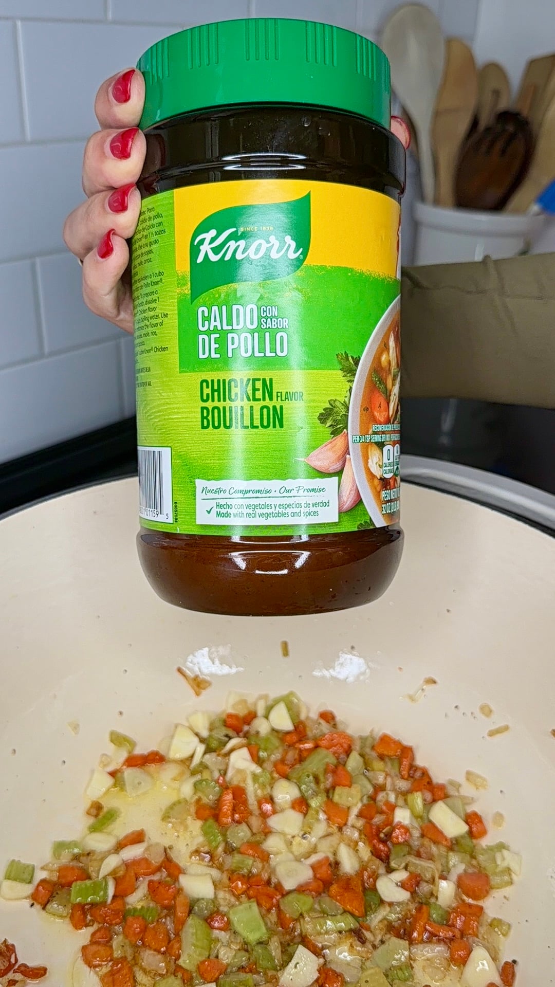 A hand holds a large jar of Knorr Caldo de Pollo chicken bouillon over a pot filled with chopped vegetables like carrots, celery, and onions, ready for cooking.